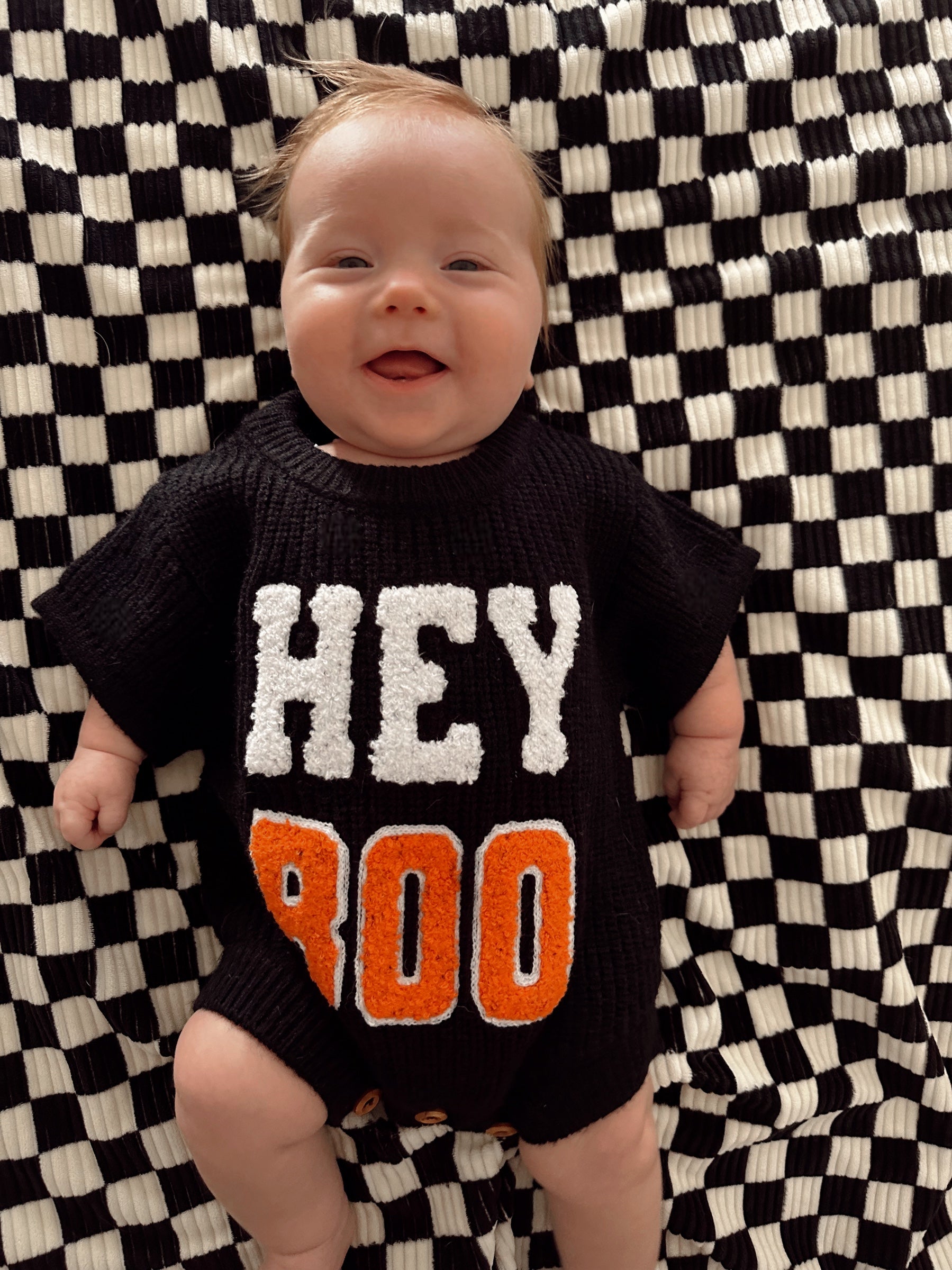 Smiling baby in a black sweater with "HEY BOO" on a checkered blanket.