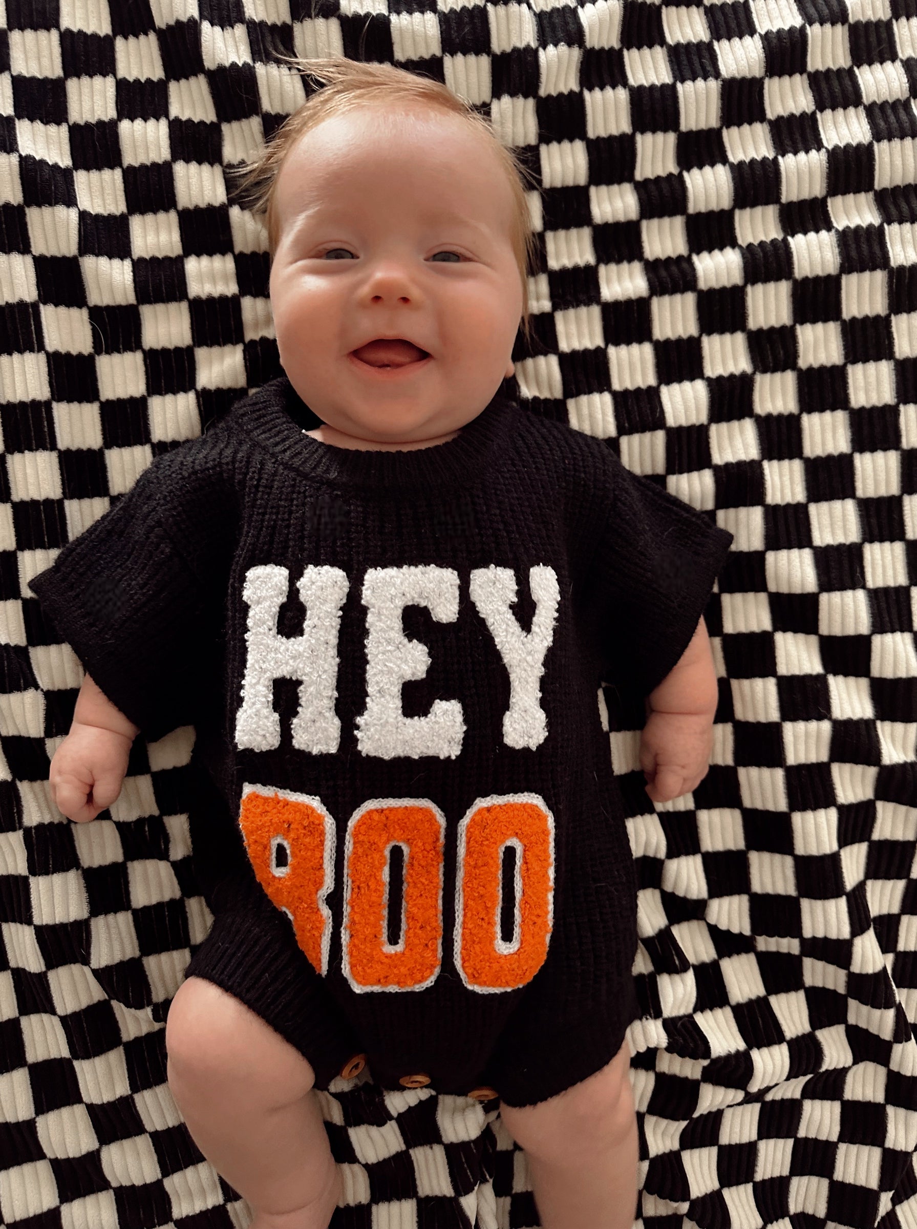 Smiling baby in a black sweater with "HEY BOO" on a checkered blanket.