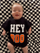 Smiling baby in a black sweater with "HEY BOO" on a checkered blanket.
