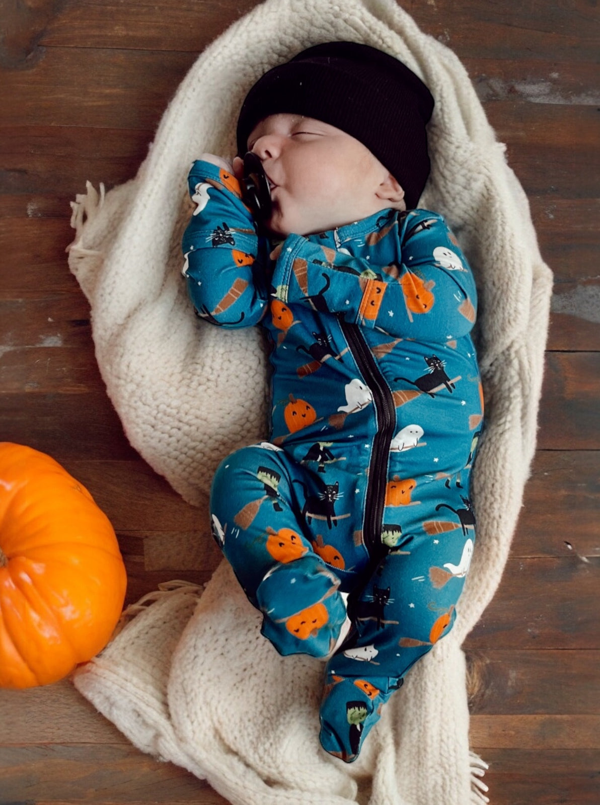Baby sleeping on a cozy blanket, wearing a blue Halloween-themed onesie, with a pumpkin nearby.