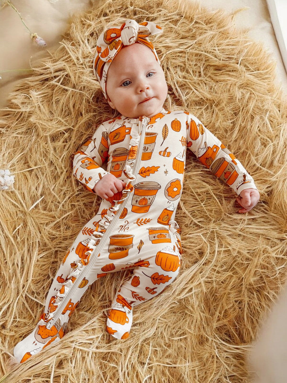 Baby in festive orange-themed outfit lying on straw, surrounded by soft textures.