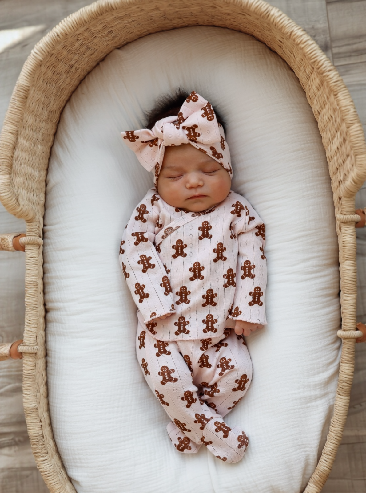 Newborn baby girl sleeping in a wicker bassinet, wearing a gingerbread-patterned outfit with a matching headband.