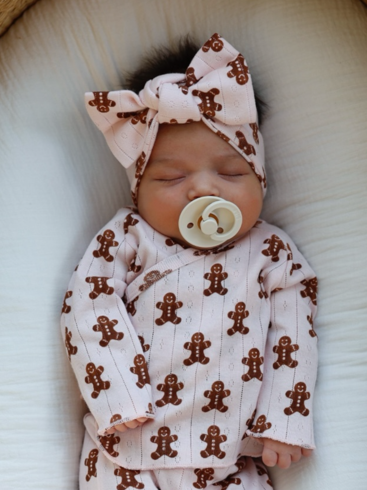 Newborn baby sleeps in pink gingerbread-patterned outfit with a matching headband and pacifier.