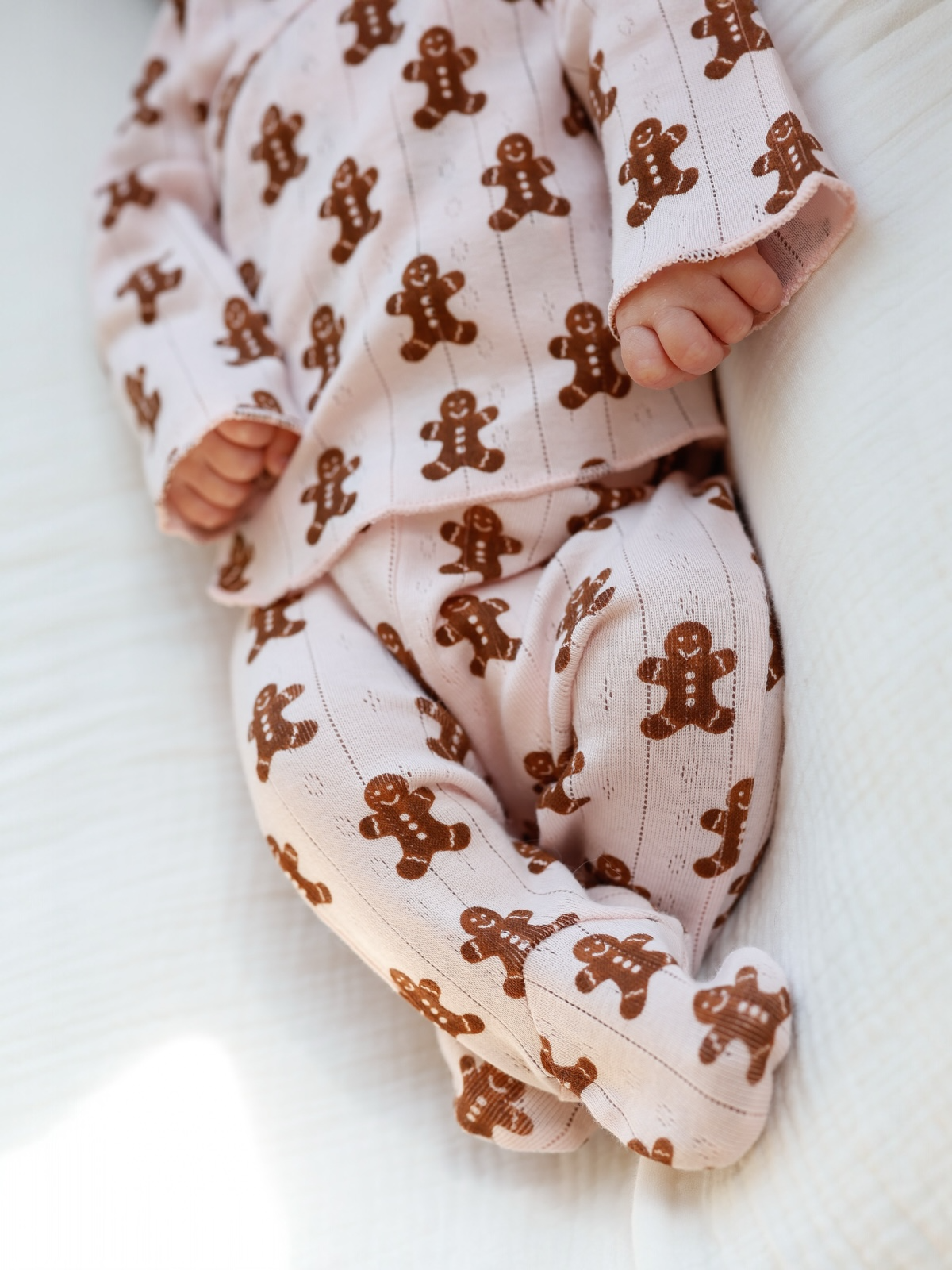 Baby in gingerbread-patterned pajamas, lying on a white blanket. Cute hands and feet visible.