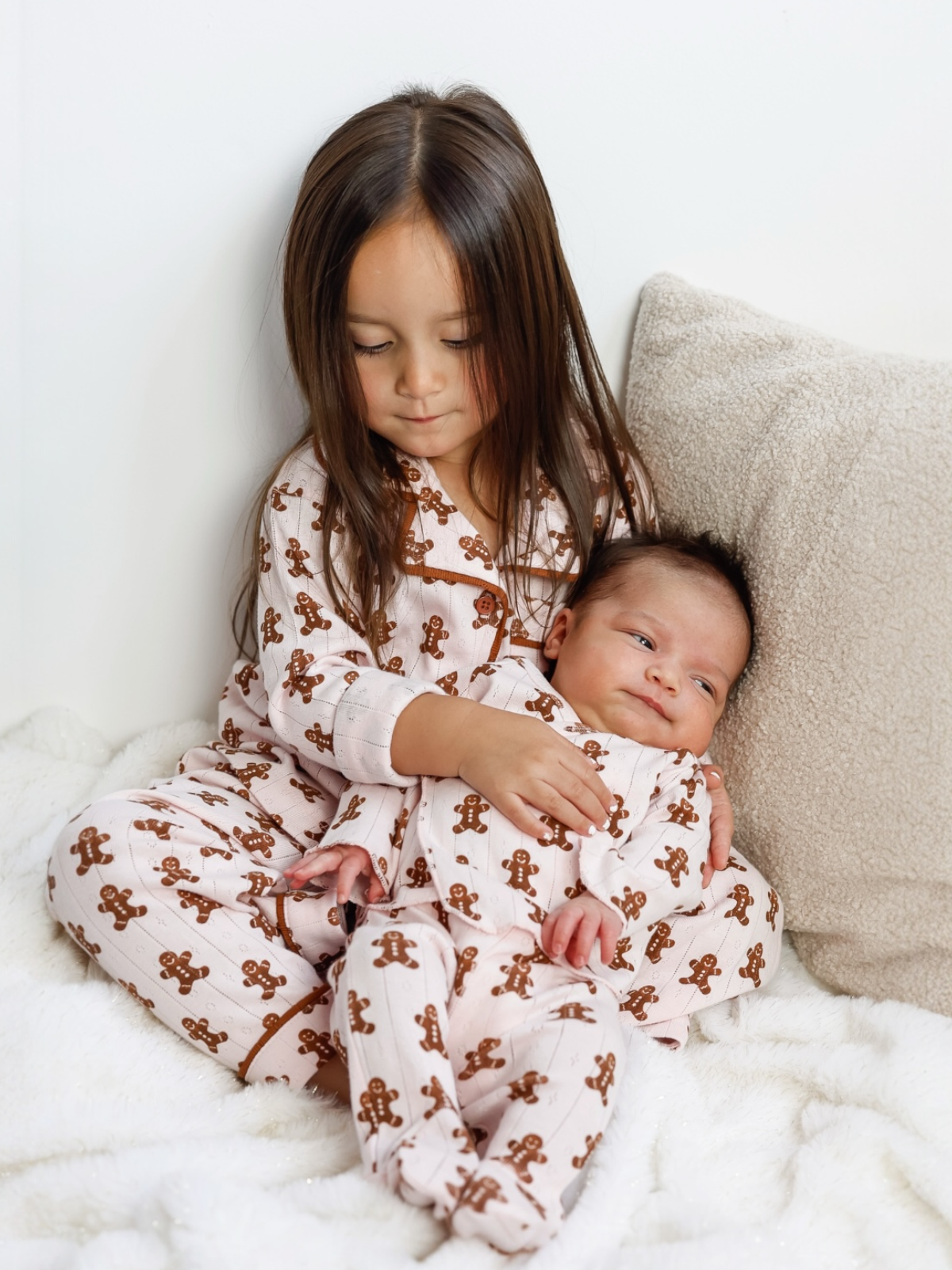 Young girl in gingerbread pajamas cuddles with a baby on a cozy blanket. They share a warm, loving moment.