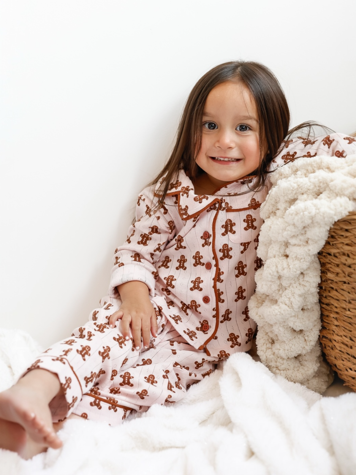 Smiling child in gingerbread pajamas, sitting on a cozy blanket next to a textured basket.
