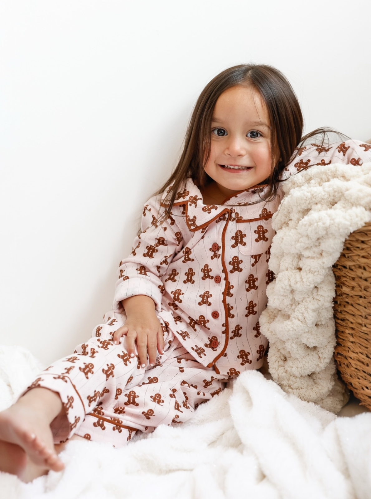 Smiling child in gingerbread pajamas, sitting on a cozy blanket next to a textured basket.