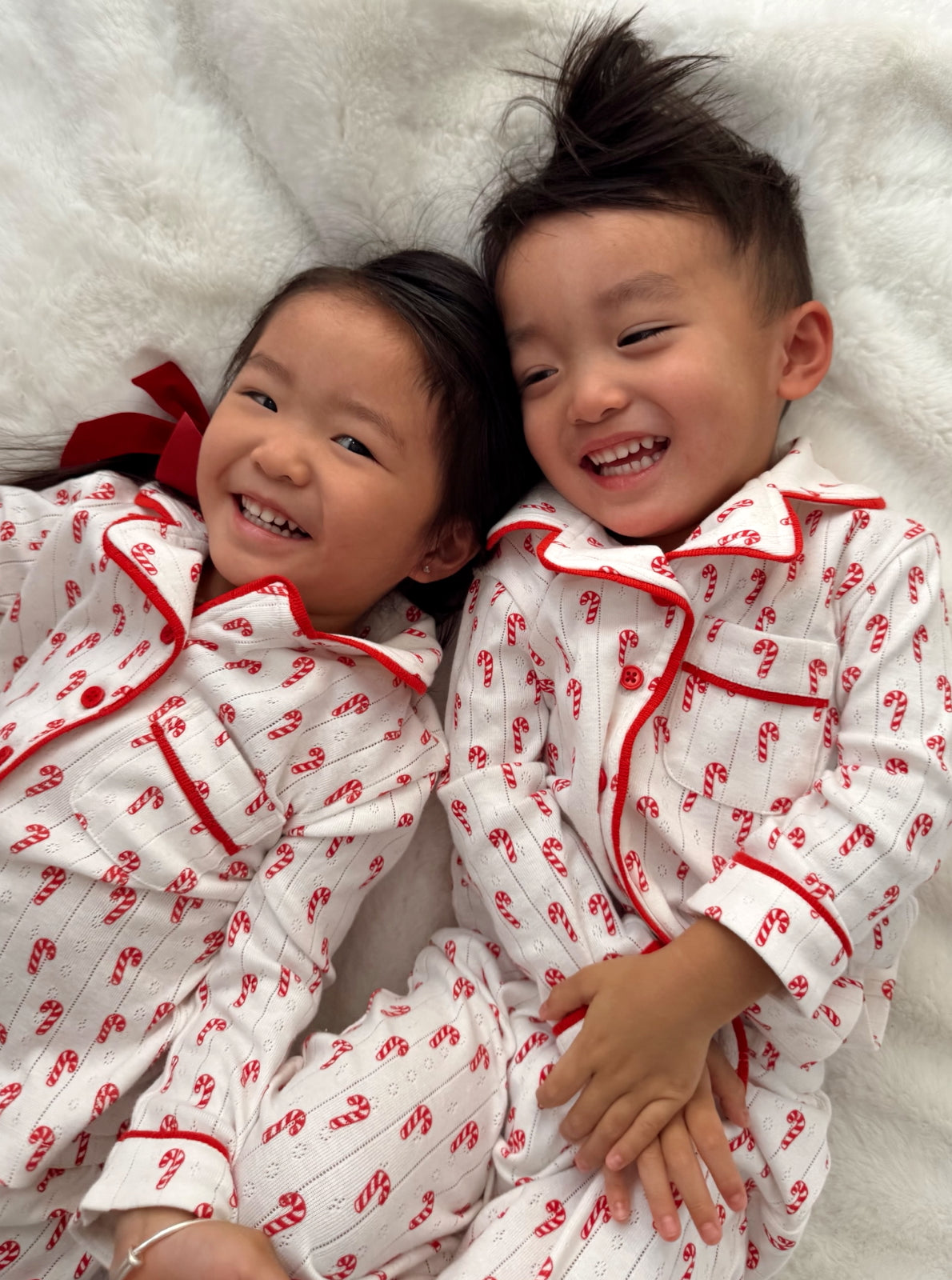 Two smiling children in festive pajamas with candy cane print, lying together on a soft blanket.