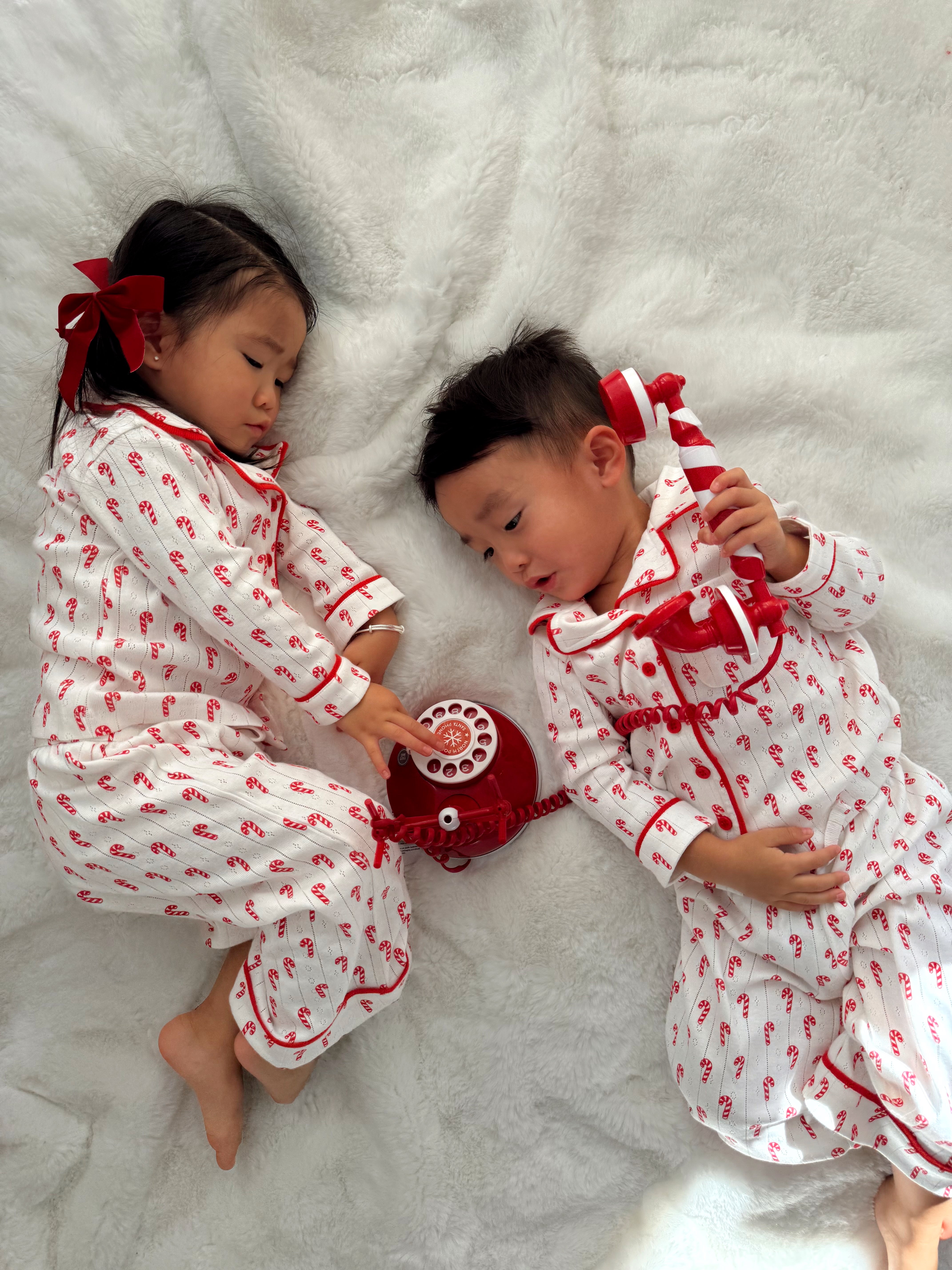 Two children in festive pajamas play with a red rotary phone on a soft white blanket.