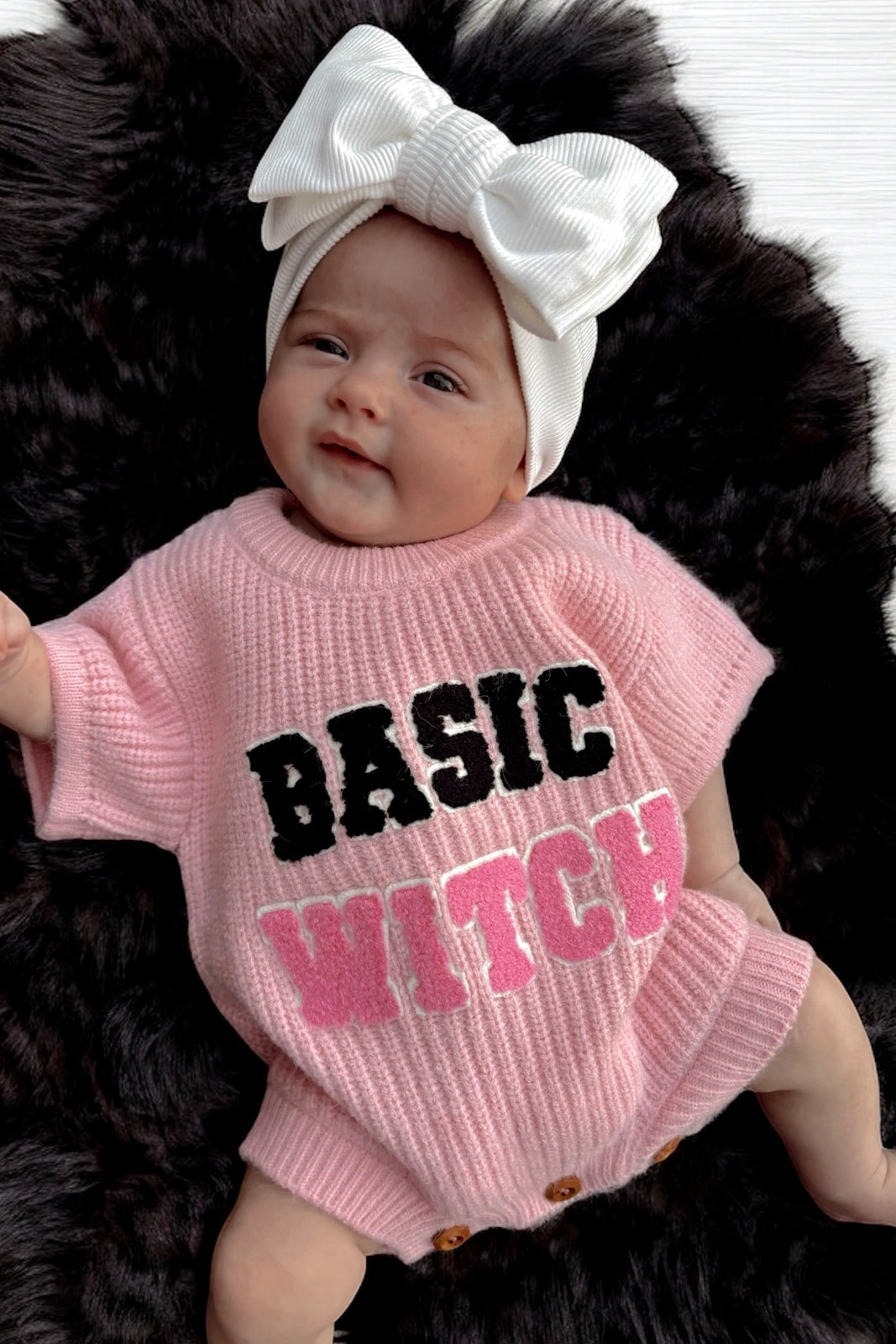 Infant wearing a pink sweater that reads "BASIC WITCH," with a large white bow on a fur background.