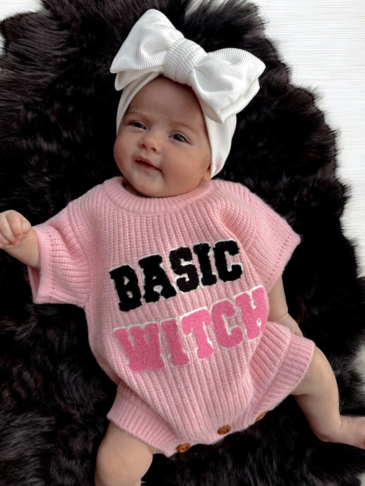 Infant wearing a pink sweater that reads "BASIC WITCH," with a large white bow on a fur background.