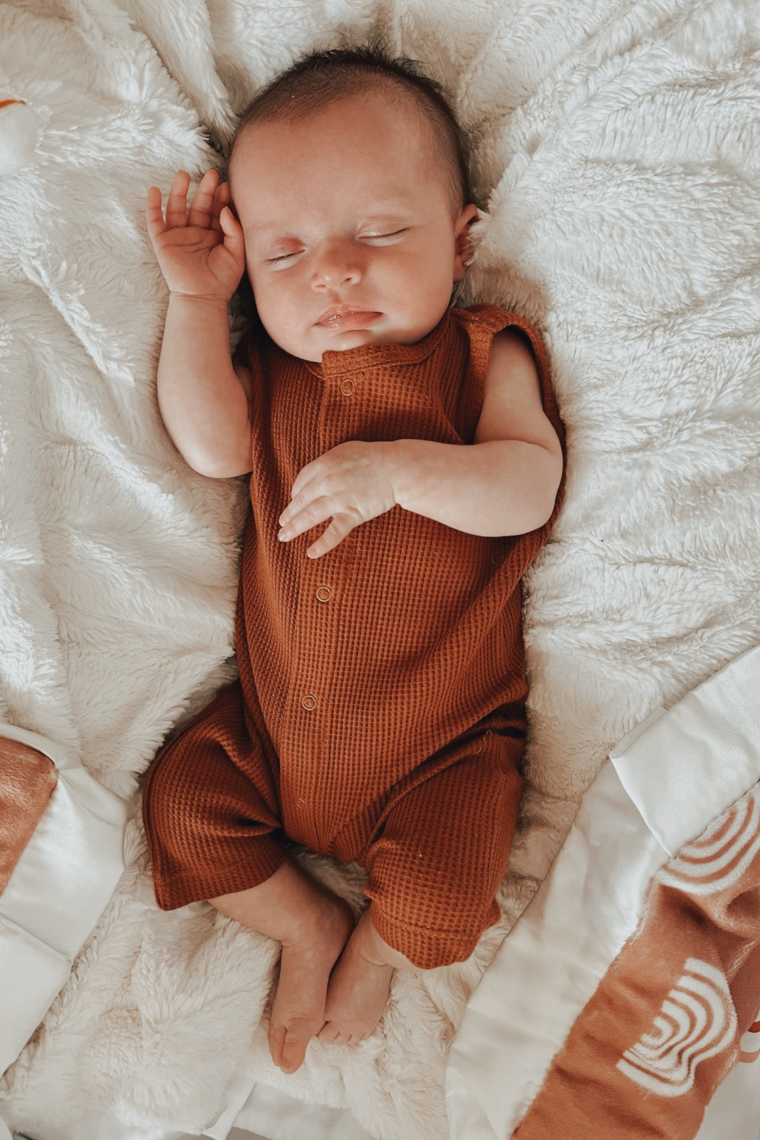 A peaceful sleeping baby in a brown outfit, resting on a fluffy white blanket.