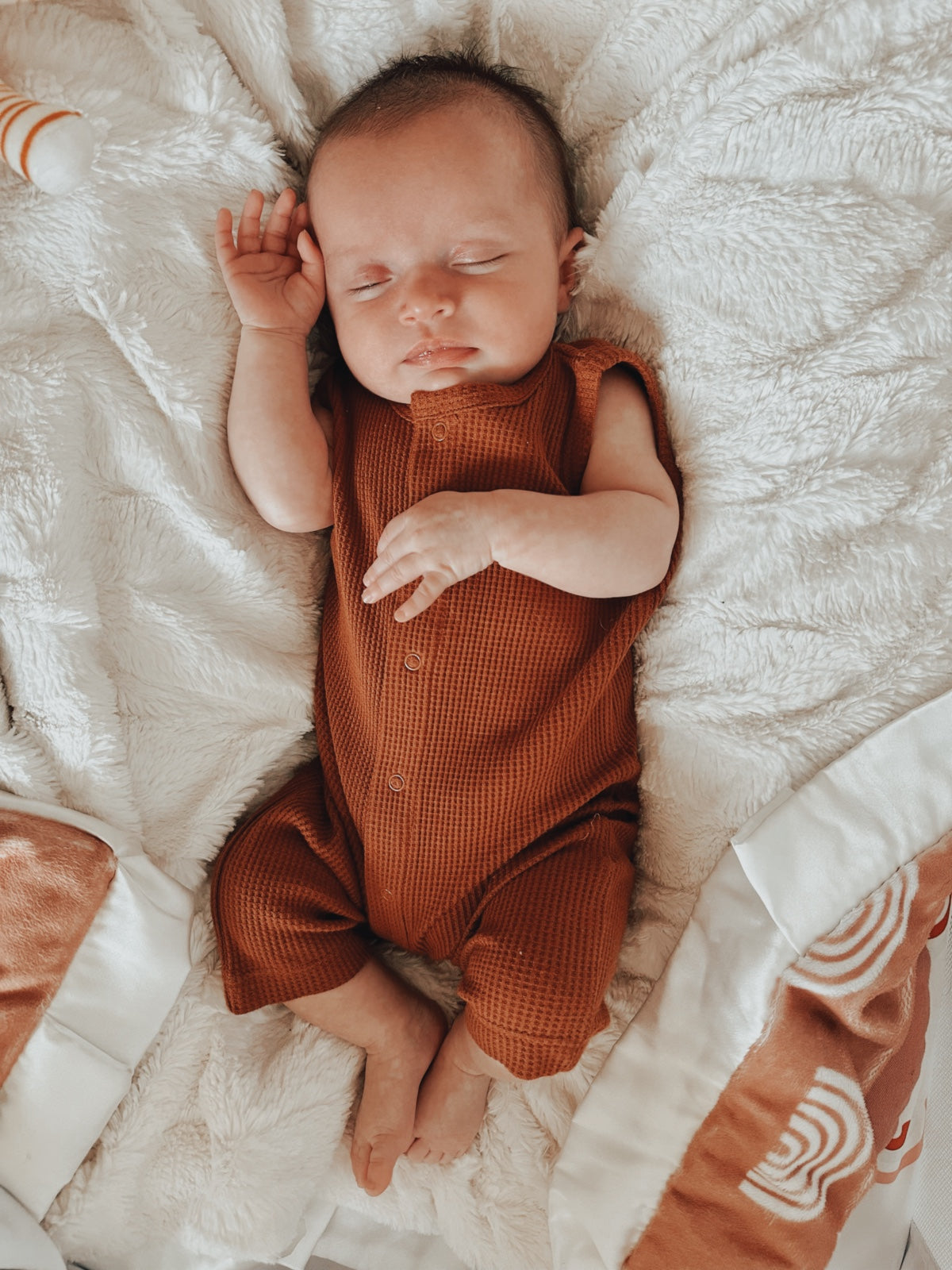 A peaceful sleeping baby in a brown outfit, resting on a fluffy white blanket.