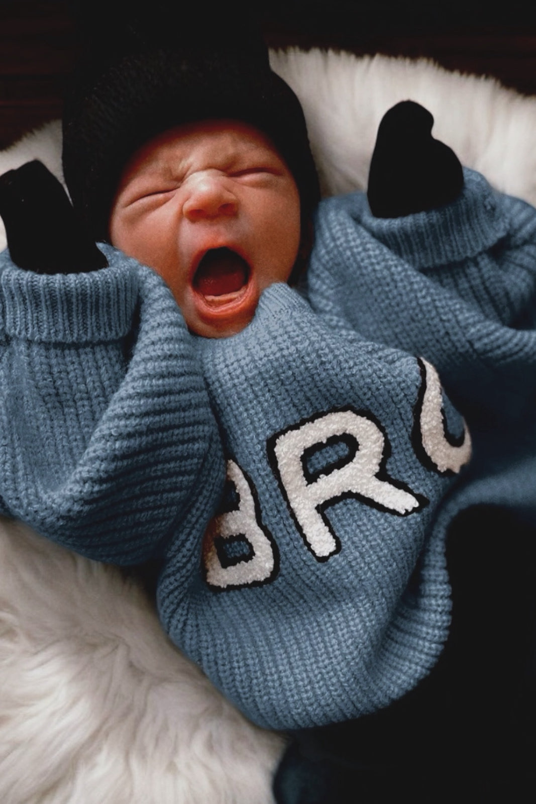 Baby in a blue sweater with "BRO" printed, yawning while lying on a fluffy white blanket.