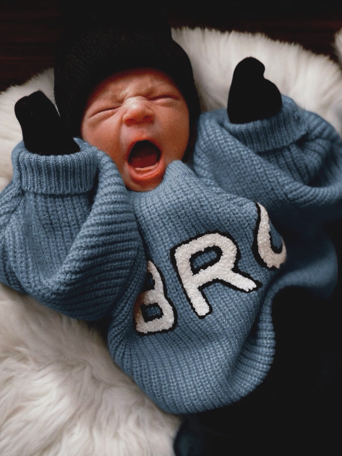 Baby in a blue sweater with "BRO" printed, yawning while lying on a fluffy white blanket.