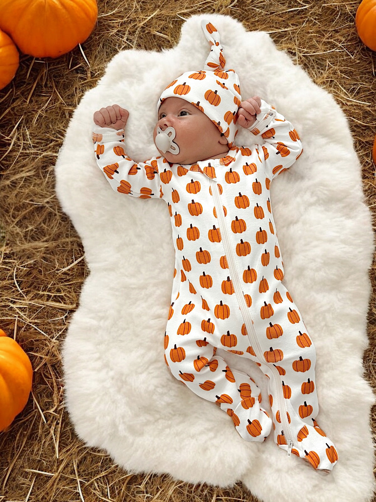 Baby in a pumpkin-patterned onesie and hat, lying on soft fabric surrounded by pumpkins.