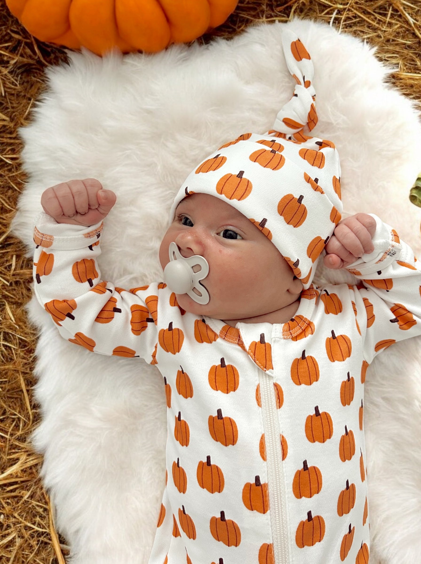 Baby in pumpkin-patterned outfit lying on a soft surface, with a pumpkin in the background.