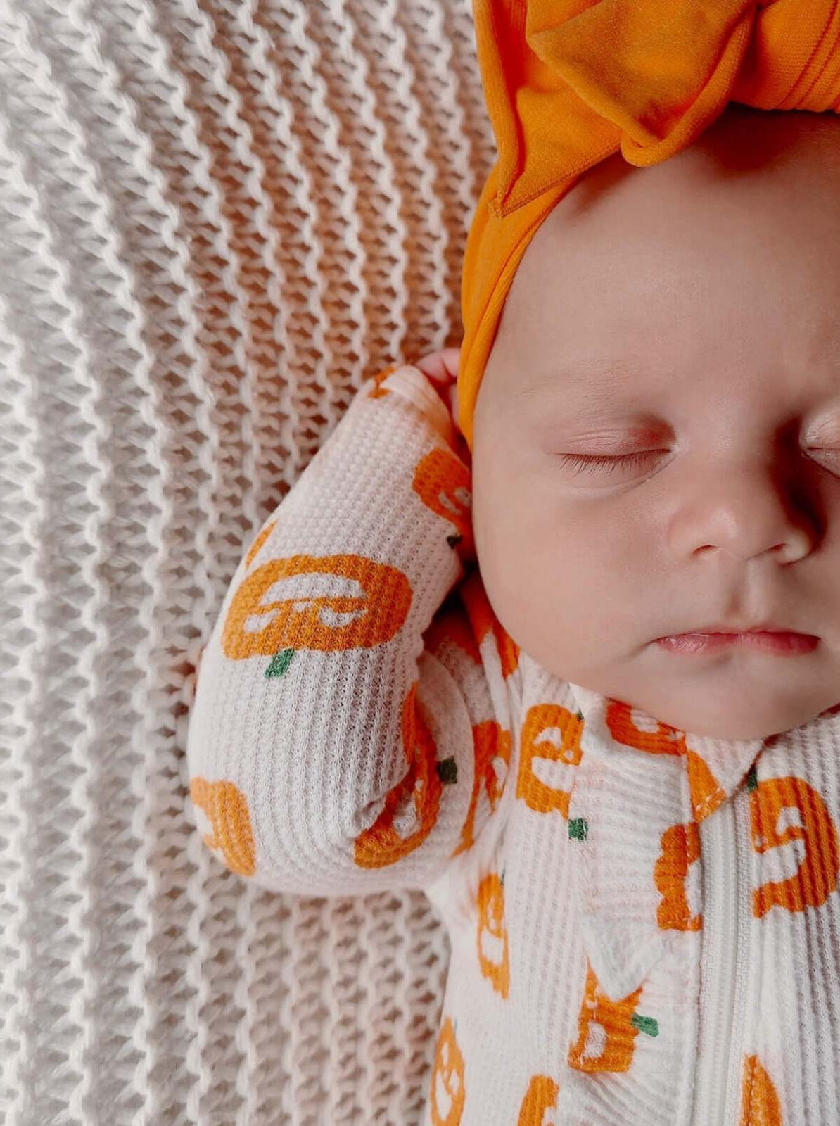 Baby wearing pumpkin-patterned outfit and orange headband, resting on a cozy textured blanket.