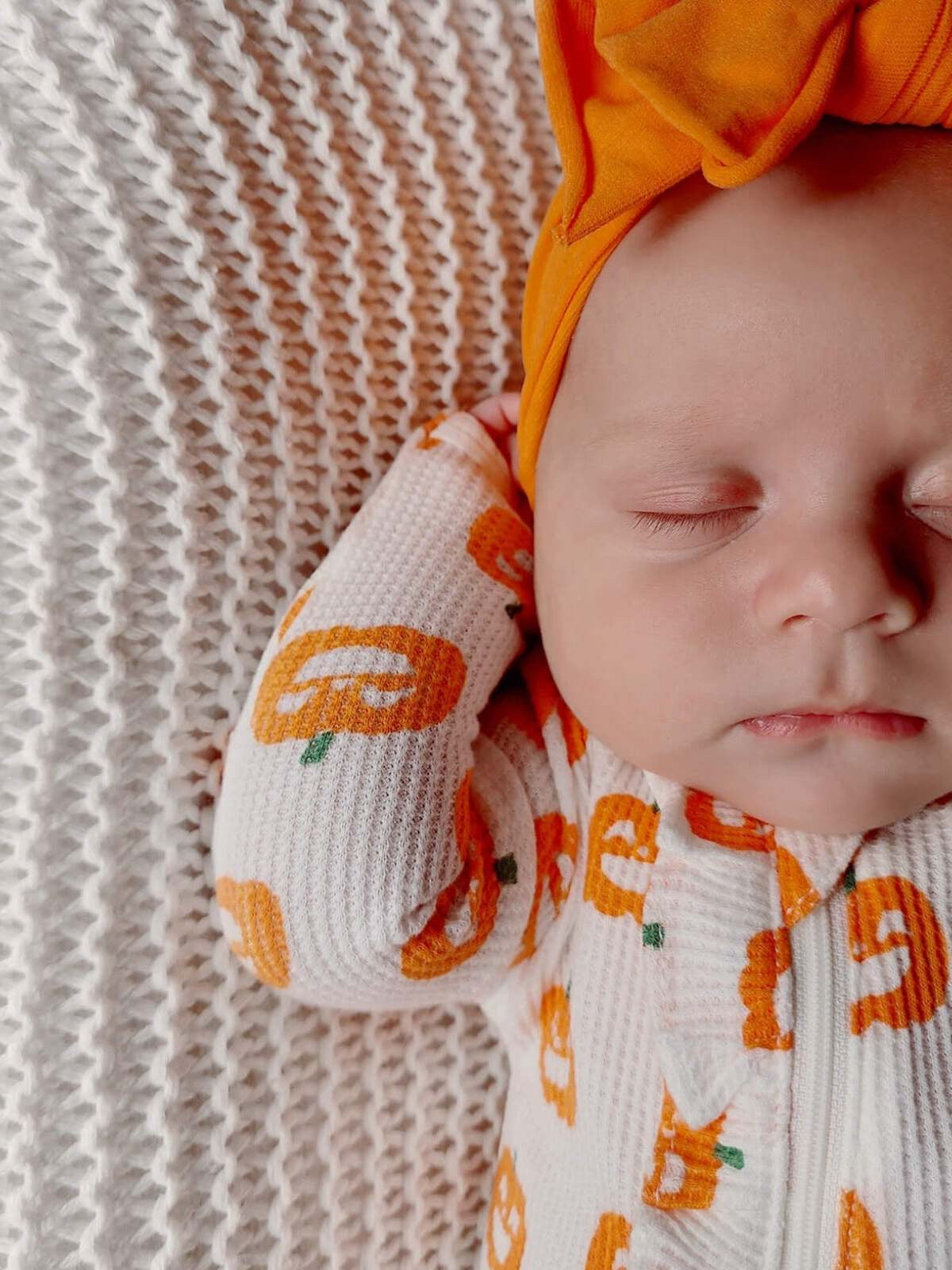 Baby wearing pumpkin-patterned outfit and orange headband, resting on a cozy textured blanket.