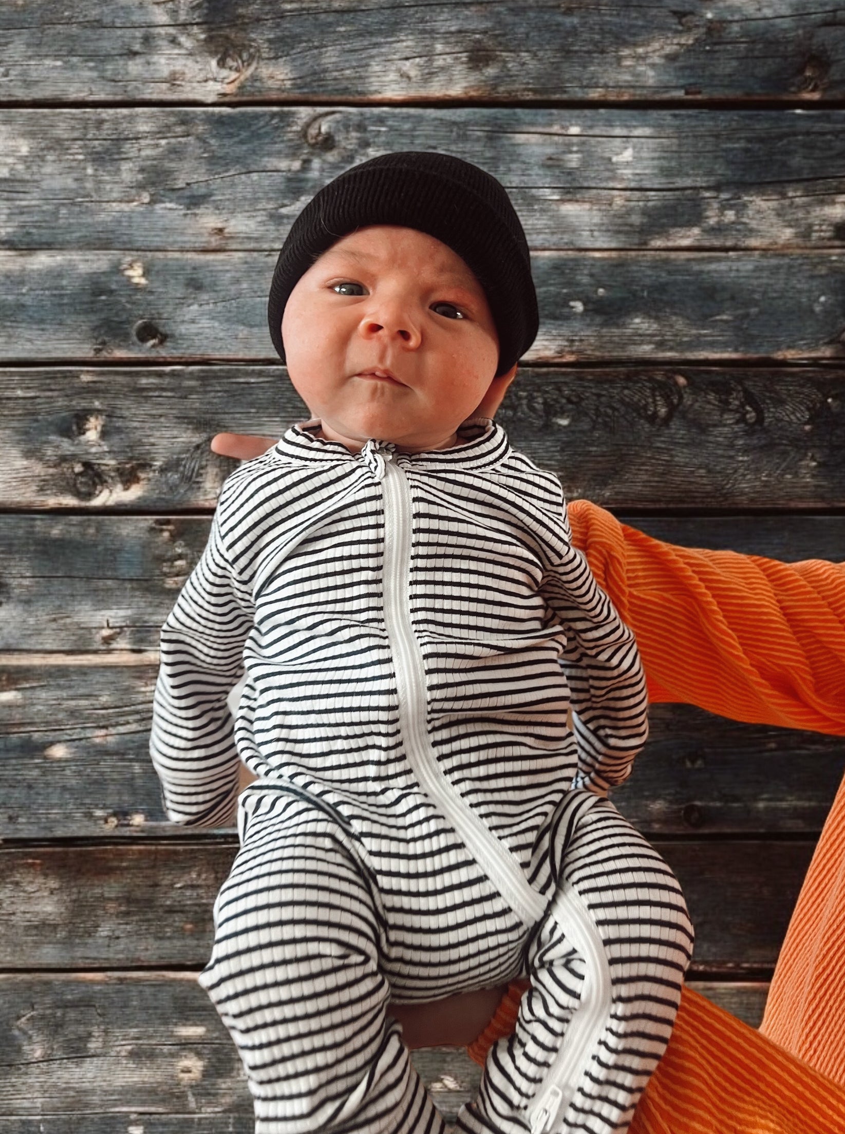 Infant in a striped onesie and black beanie held against a wooden background.