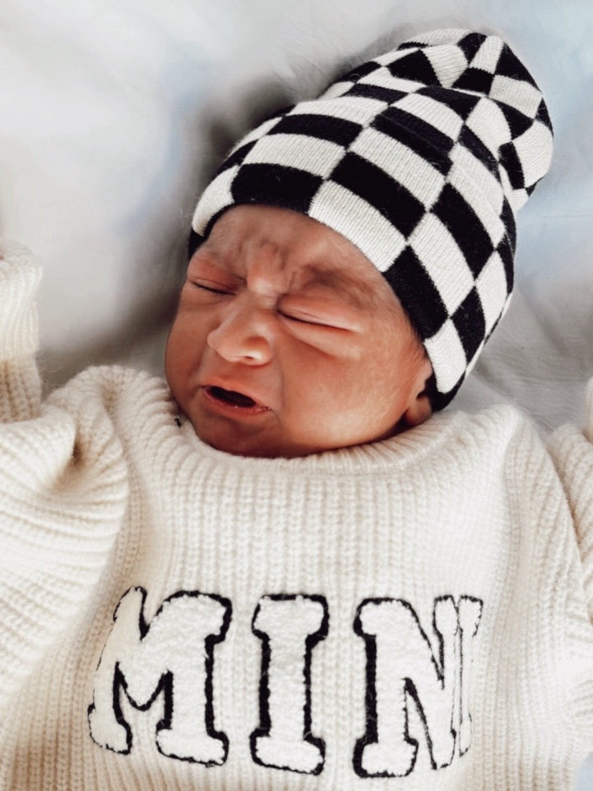 Newborn baby with a black and white checkered hat, wearing a white sweater with "MINI" printed on it, crying.