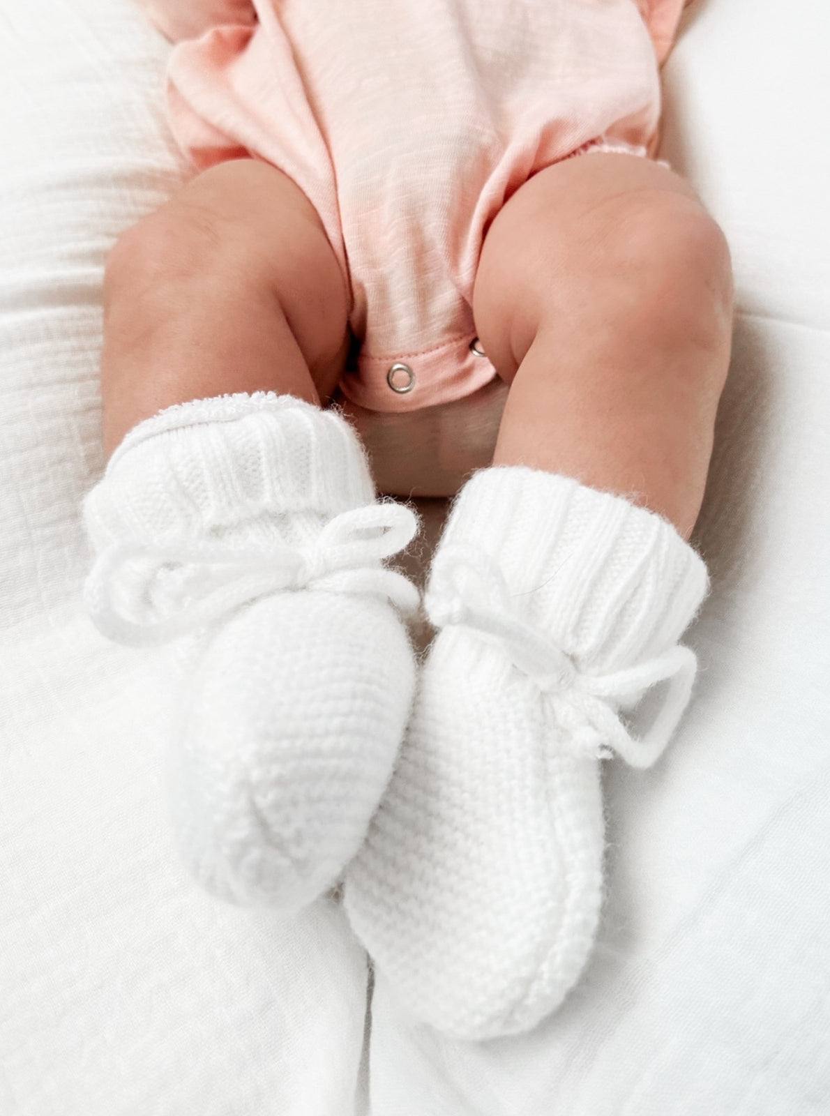 Baby feet in soft white knitted booties, resting on a soft white blanket, wearing a light pink outfit.
