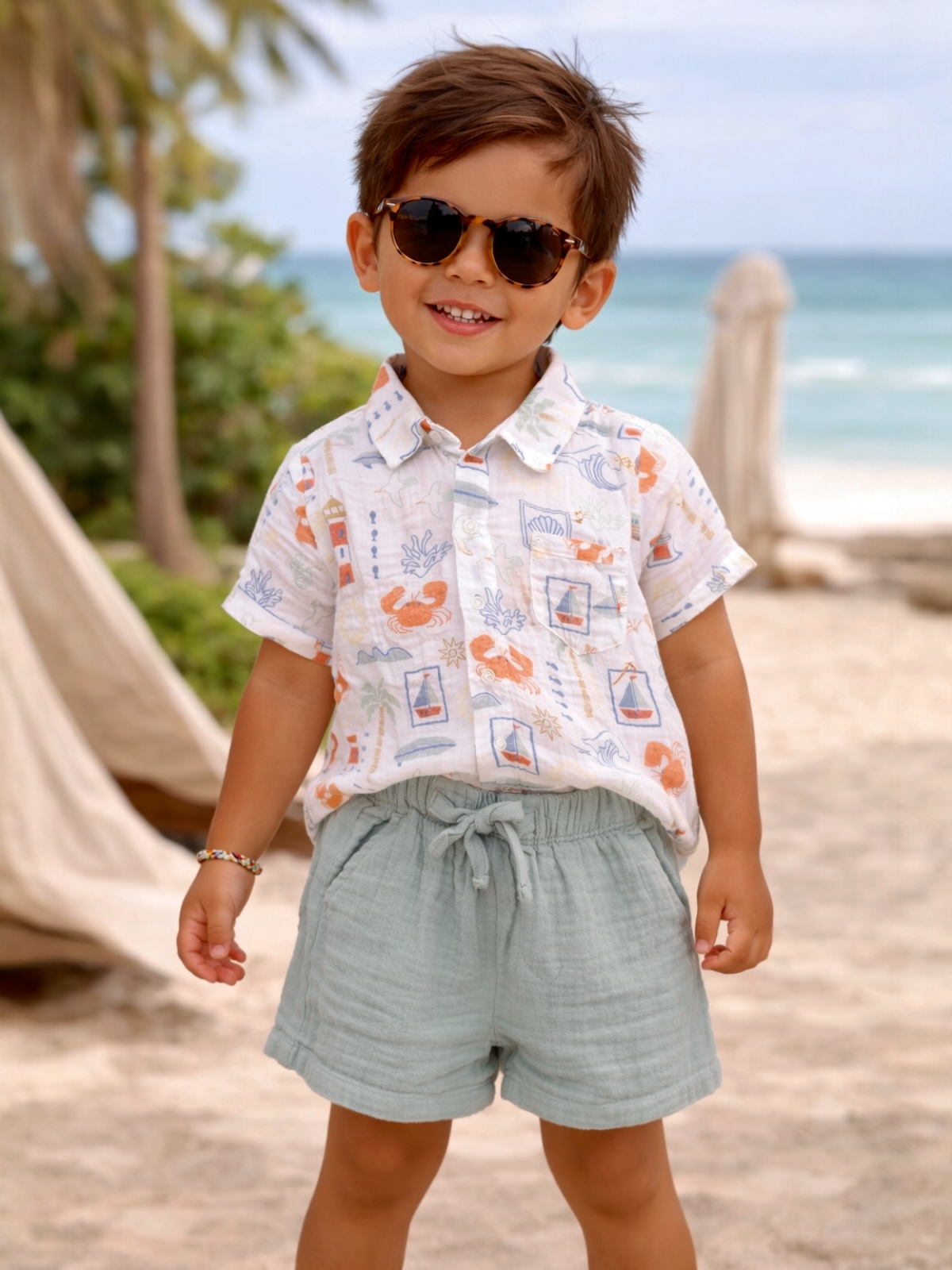 Smiling child in sunglasses and a colorful shirt stands on the beach, with ocean and palm trees in the background.