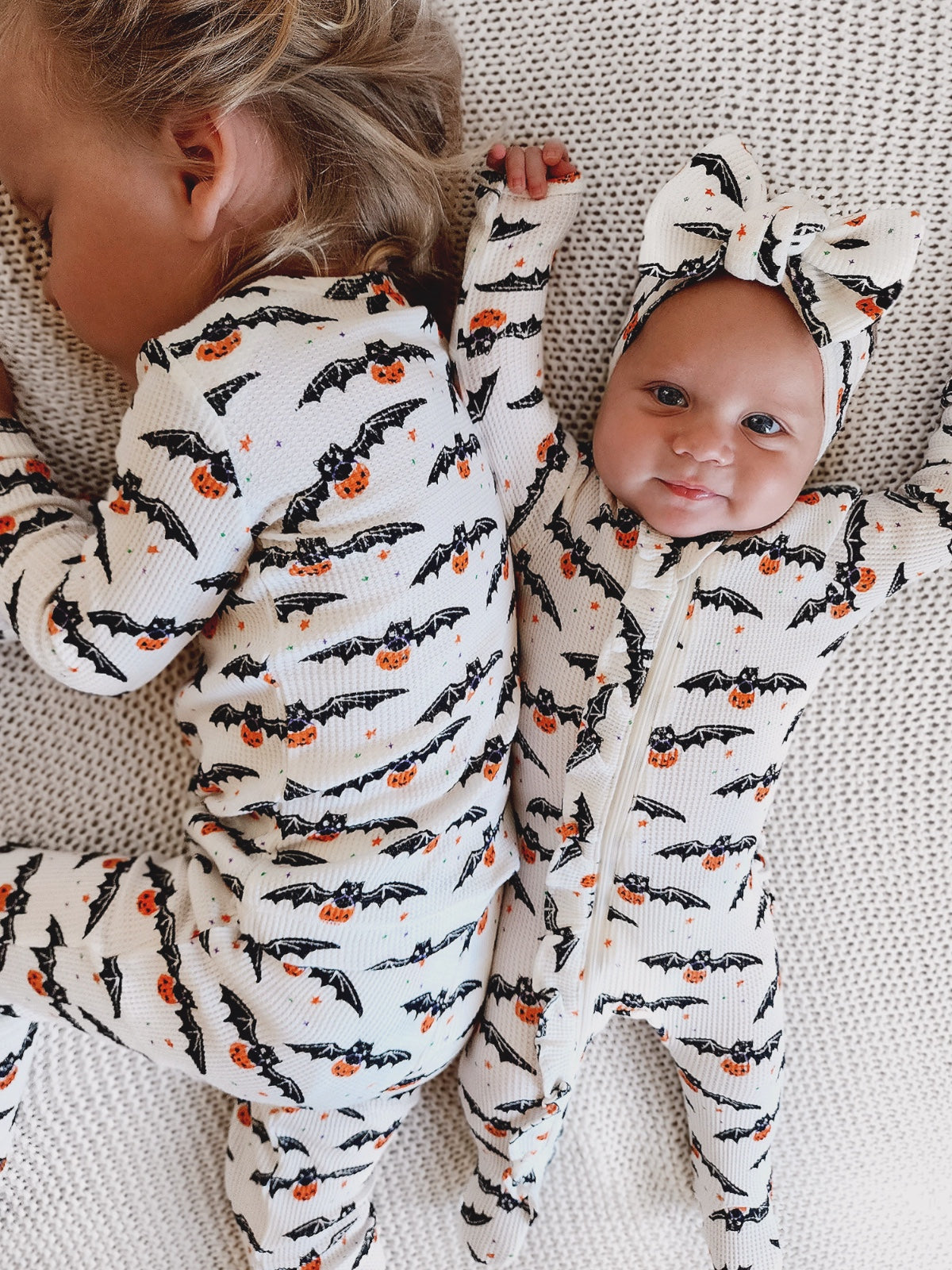 Two siblings in Halloween-themed pajamas with bats and pumpkins, posed on a cozy blanket.