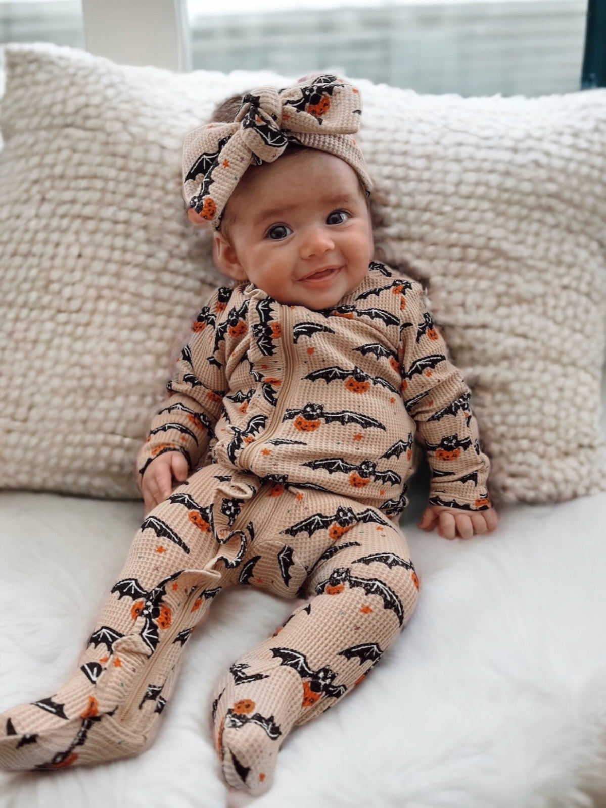 Smiling baby in a bat and pumpkin outfit sits on a cozy blanket, wearing a matching headband.
