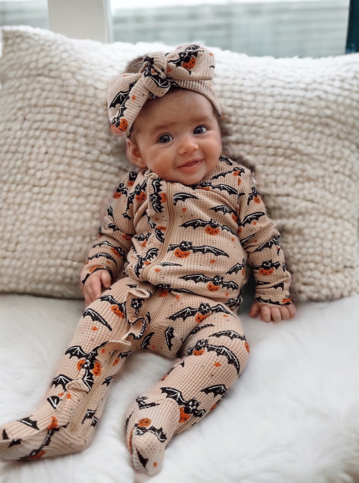 Smiling baby in a bat and pumpkin outfit sits on a cozy blanket, wearing a matching headband.