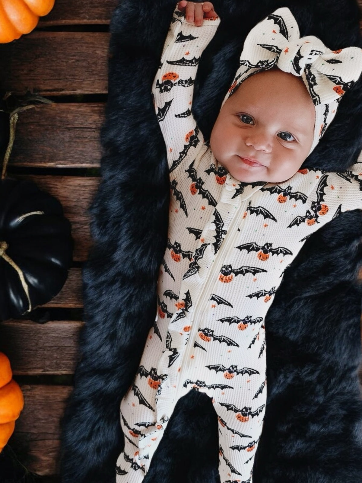 Baby in a bat-patterned onesie and bow, lying on black fur with pumpkins and decorative gourds around.