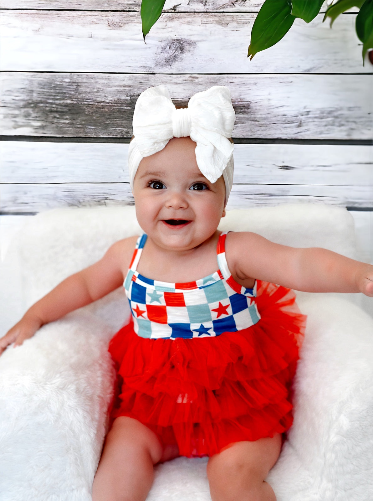 Smiling baby in a red ruffled outfit with a patterned top and large white bow on a fluffy chair against a wooden backdrop.