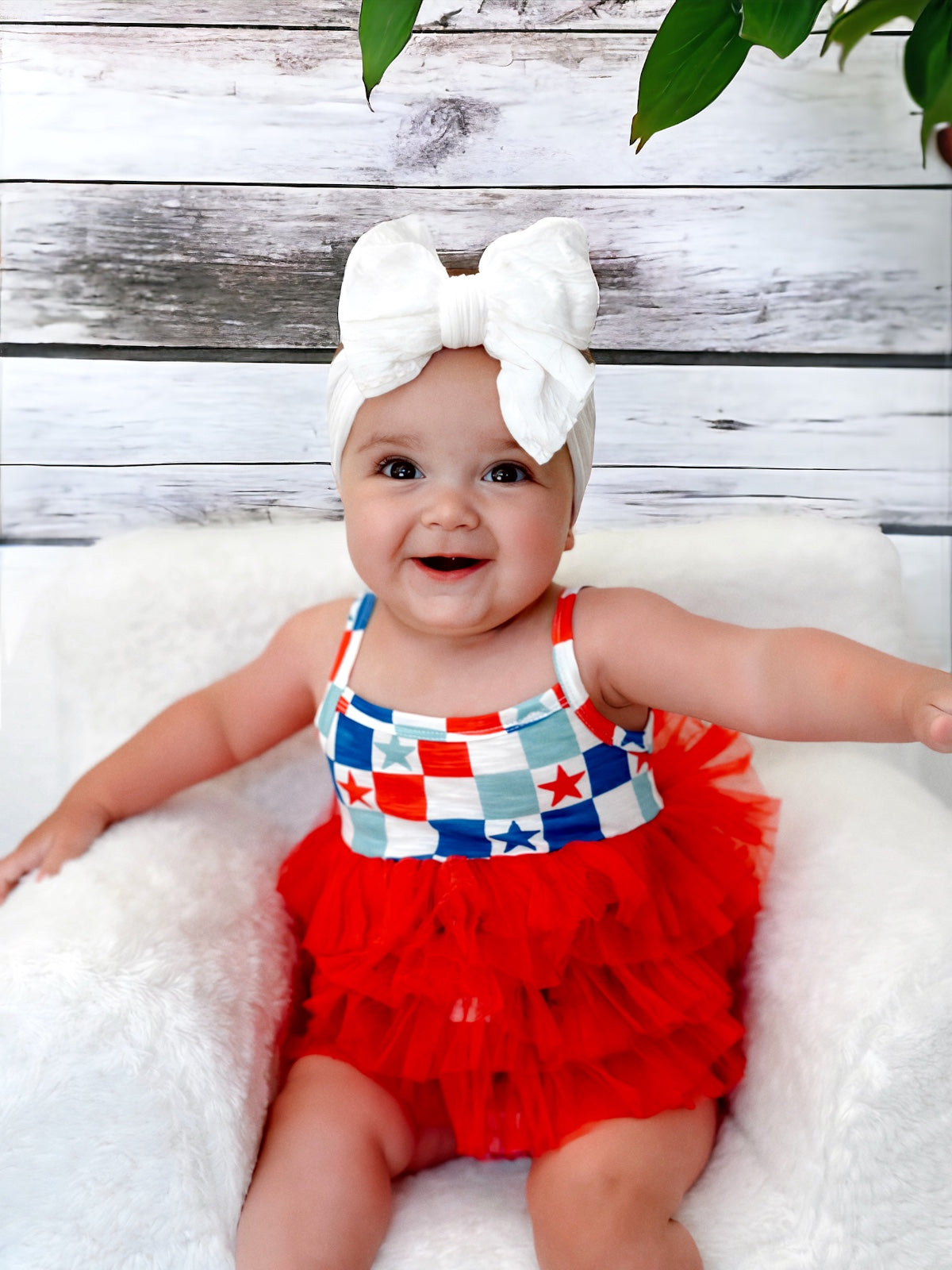 Smiling baby in a red ruffled outfit with a patterned top and large white bow on a fluffy chair against a wooden backdrop.