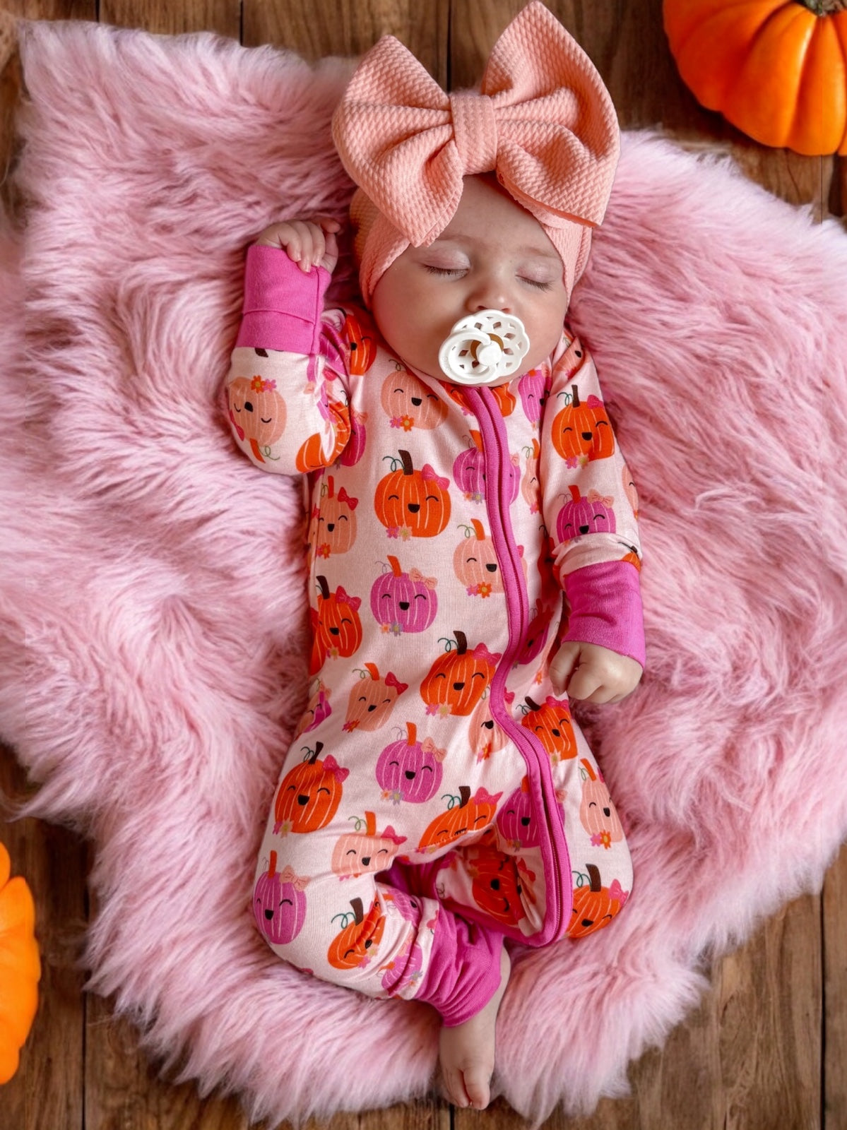 Baby wearing a pumpkin-patterned onesie, asleep on a pink fluffy blanket, with a large pink bow headband nearby.