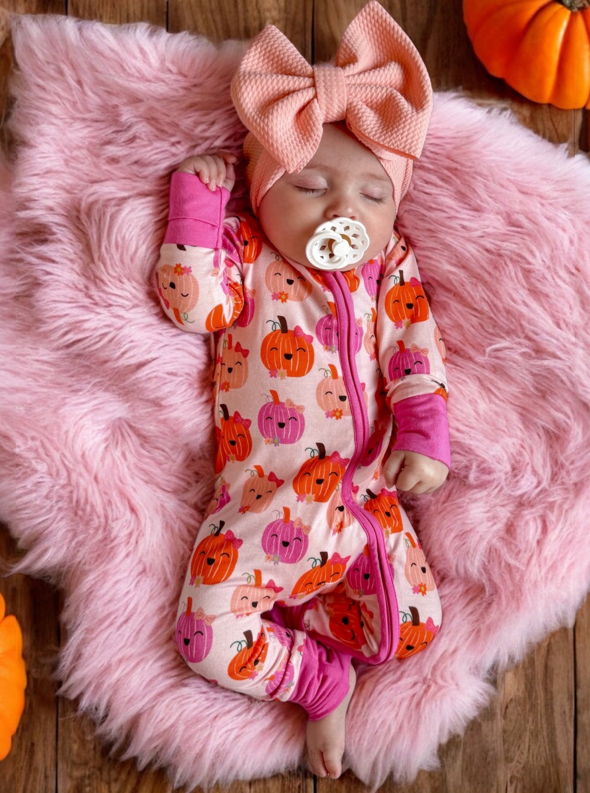 Baby wearing a pumpkin-patterned onesie, asleep on a pink fluffy blanket, with a large pink bow headband nearby.