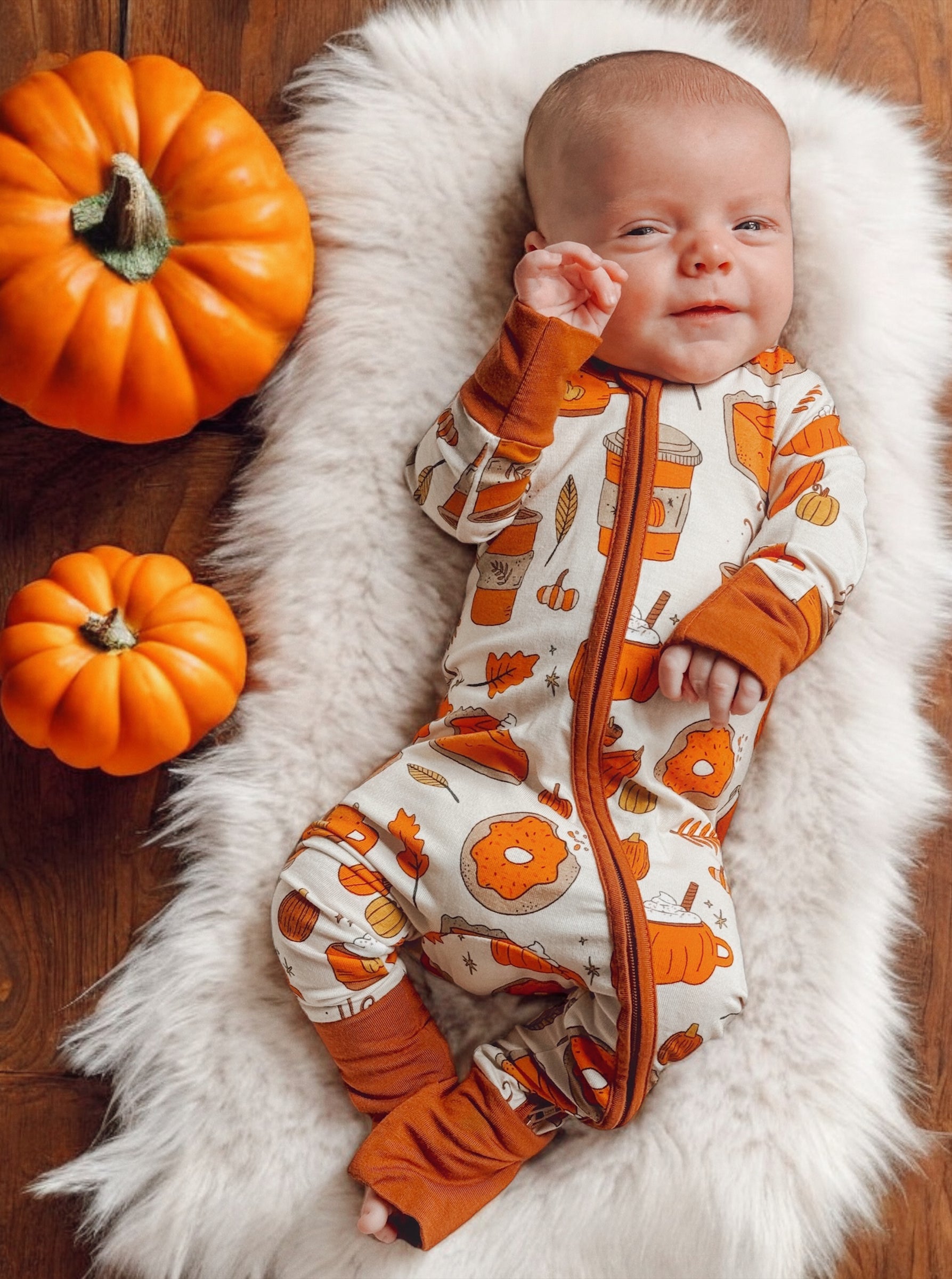 Baby in pumpkin-themed outfit on a fluffy blanket, surrounded by pumpkins, smiling and waving.