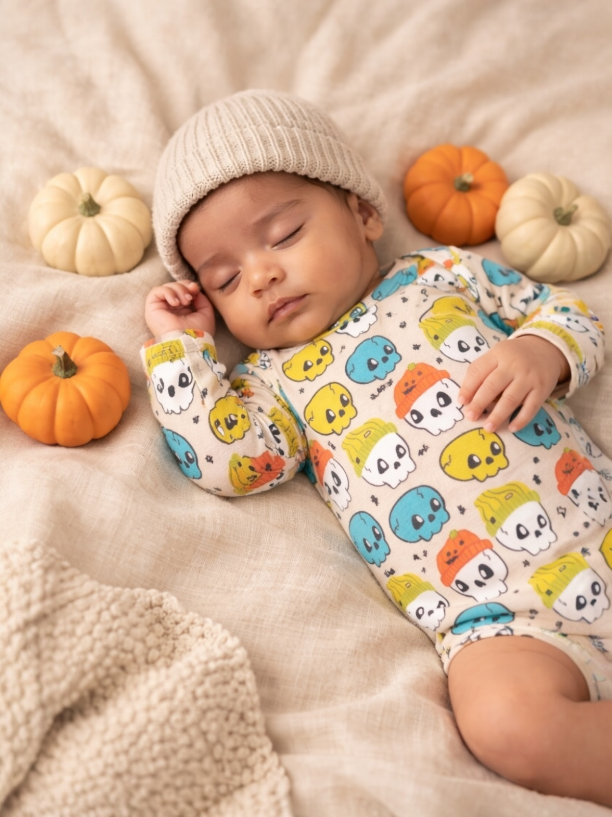 Sleeping baby in a skull-patterned onesie surrounded by small pumpkins on a soft, textured blanket.