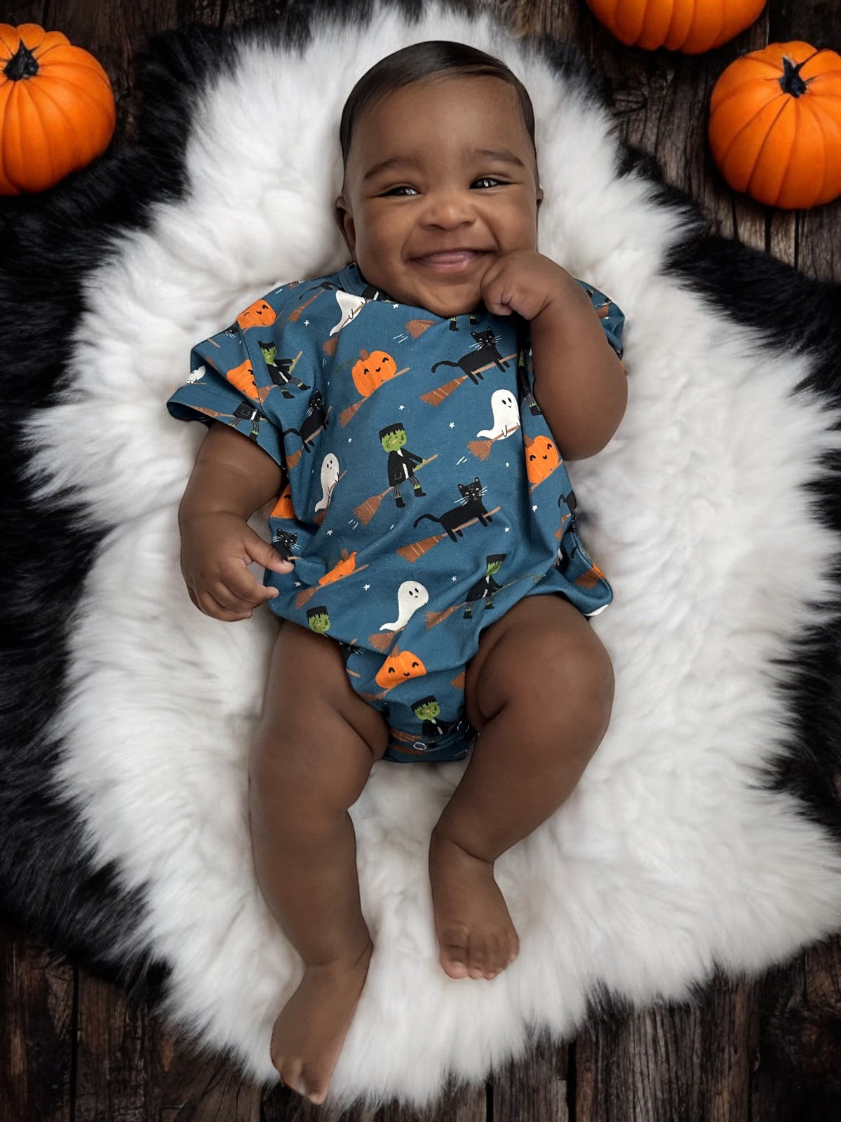 Smiling baby in a cute Halloween-themed outfit, surrounded by small pumpkins on a fluffy white rug.