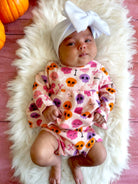 Infant girl in a festive skull-patterned outfit and white bow on a fluffy blanket, surrounded by orange pumpkins.
