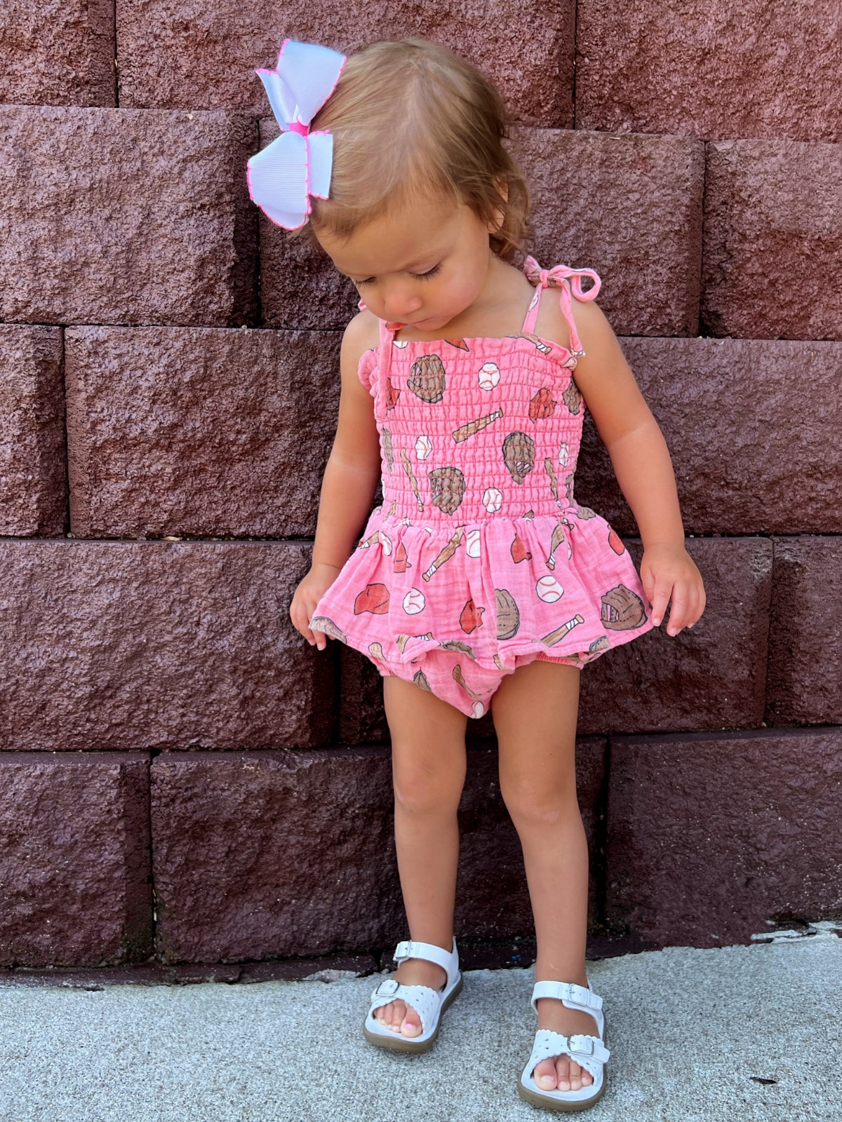 Toddler girl in a pink outfit with ice cream prints, standing on a sidewalk in front of a textured brick wall.
