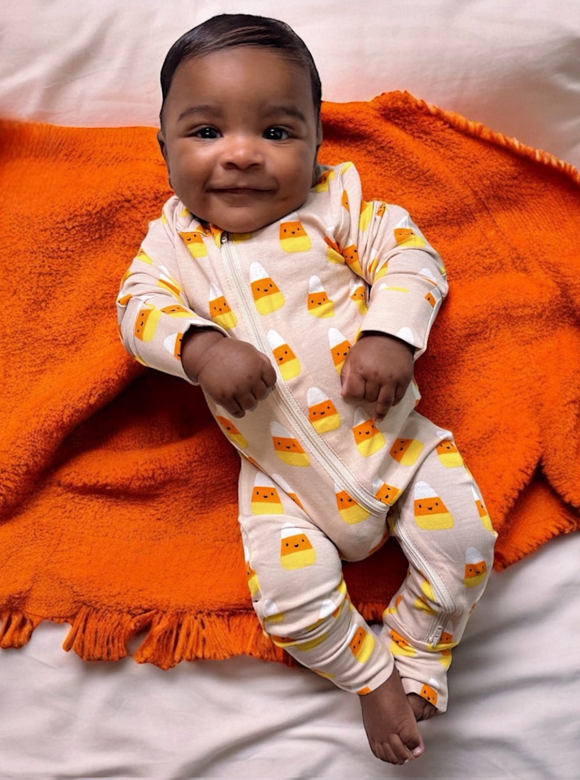 Smiling baby in candy corn pajamas on an orange blanket, relaxed and happy.