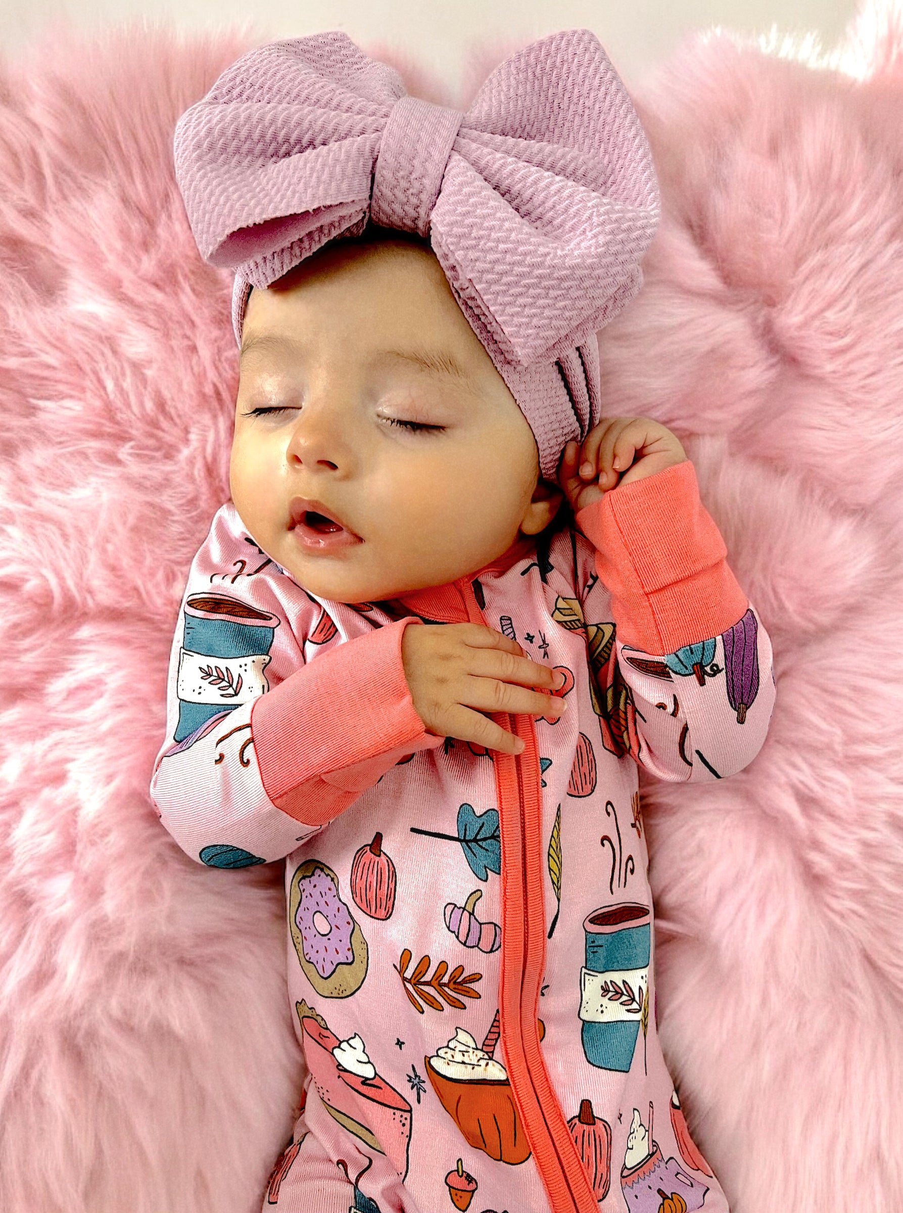 Infant girl in playful pajamas and a large bow, resting on pink fluffy fabric.