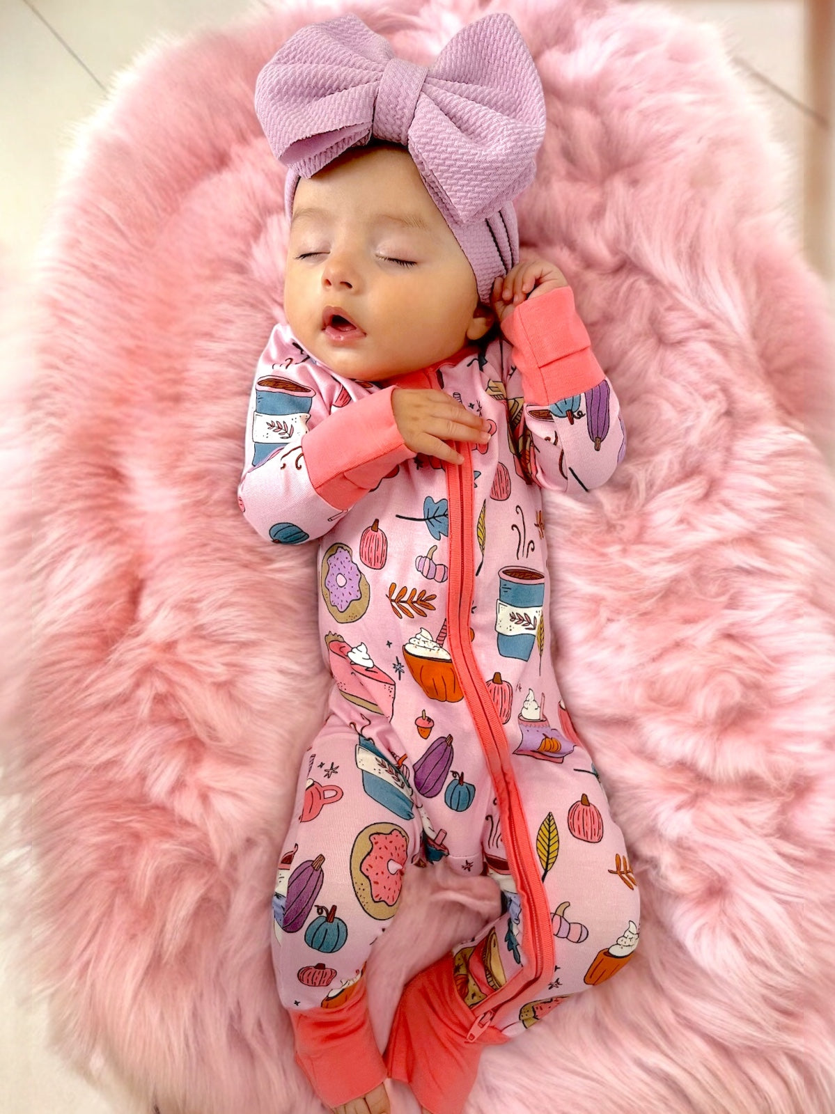Baby sleeping on a pink furry blanket, wearing a patterned onesie and a large bow headband.