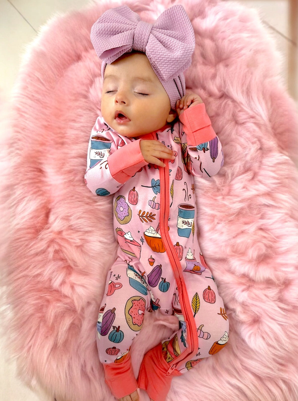 Baby sleeping on a pink furry blanket, wearing a patterned onesie and a large bow headband.