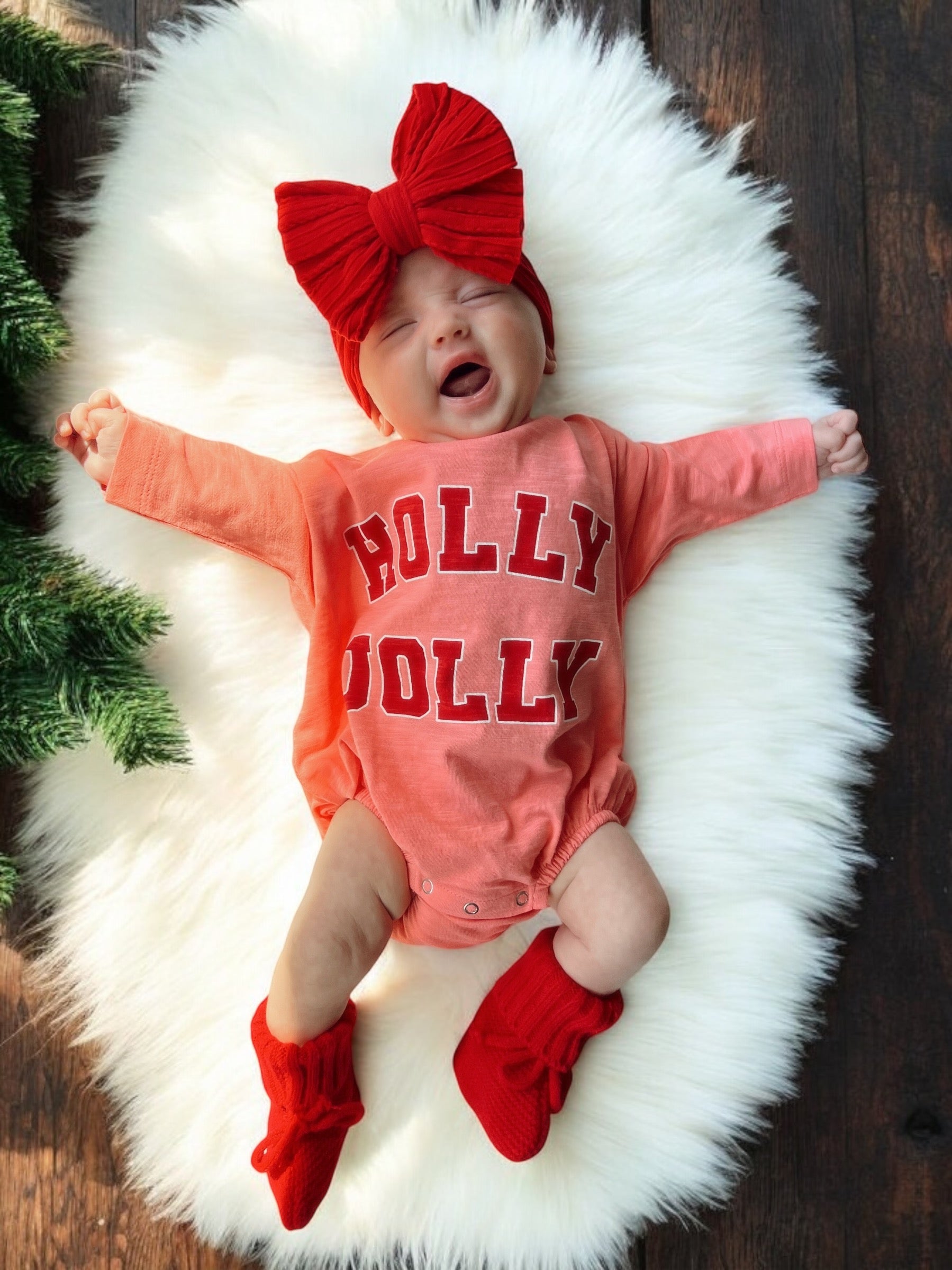Smiling baby on a fluffy rug, wearing a red bow and a "HOLLY JOLLY" outfit, with festive decorations nearby.