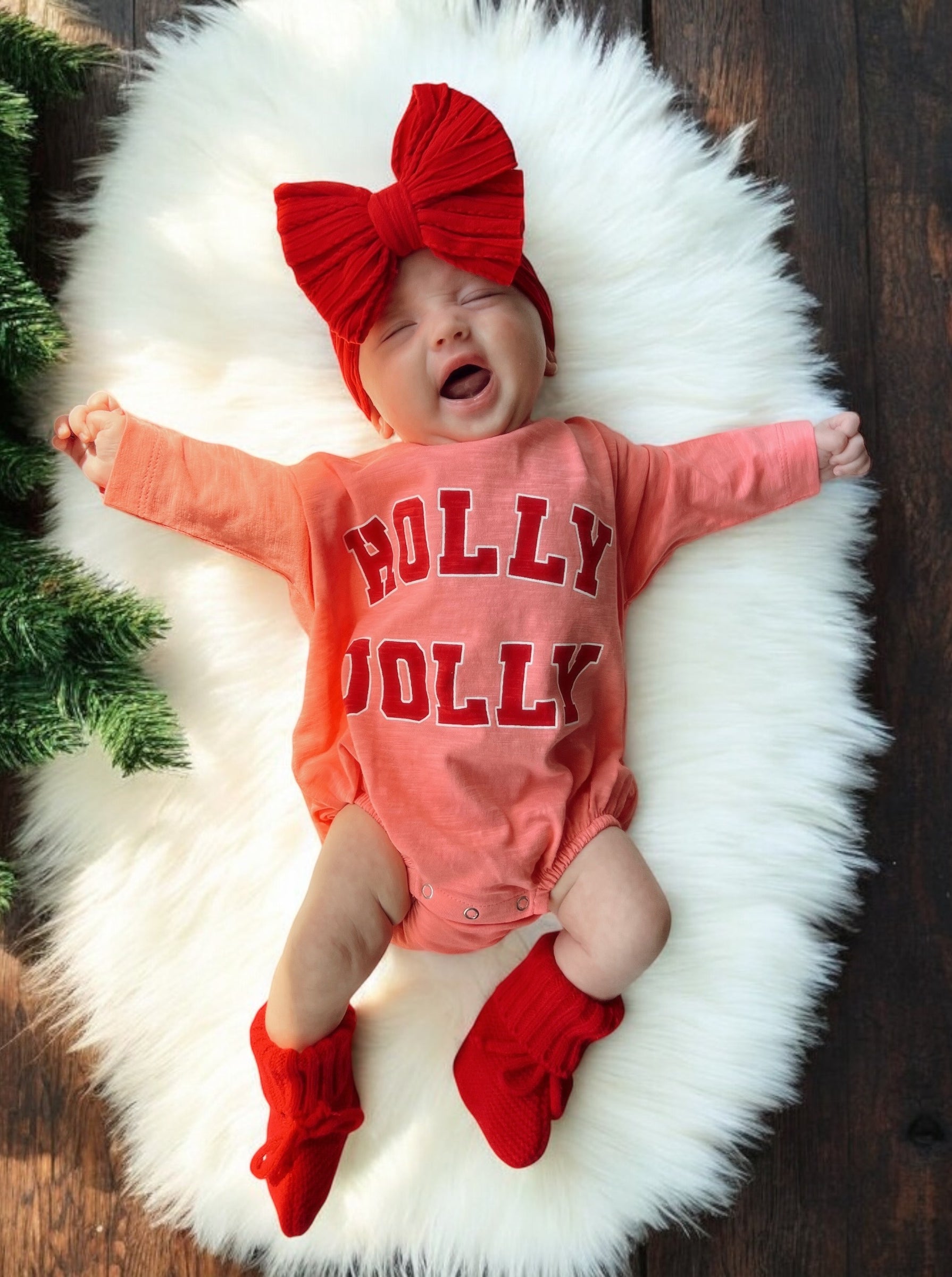 Smiling baby on a fluffy rug, wearing a red bow and a "HOLLY JOLLY" outfit, with festive decorations nearby.