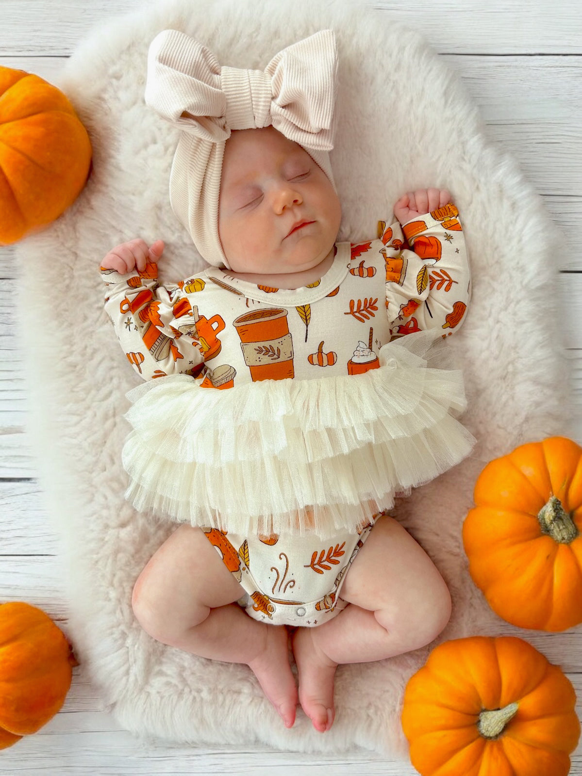 Baby girl in festive outfit with pumpkins, sleeping on a fluffy mat, surrounded by small orange pumpkins.