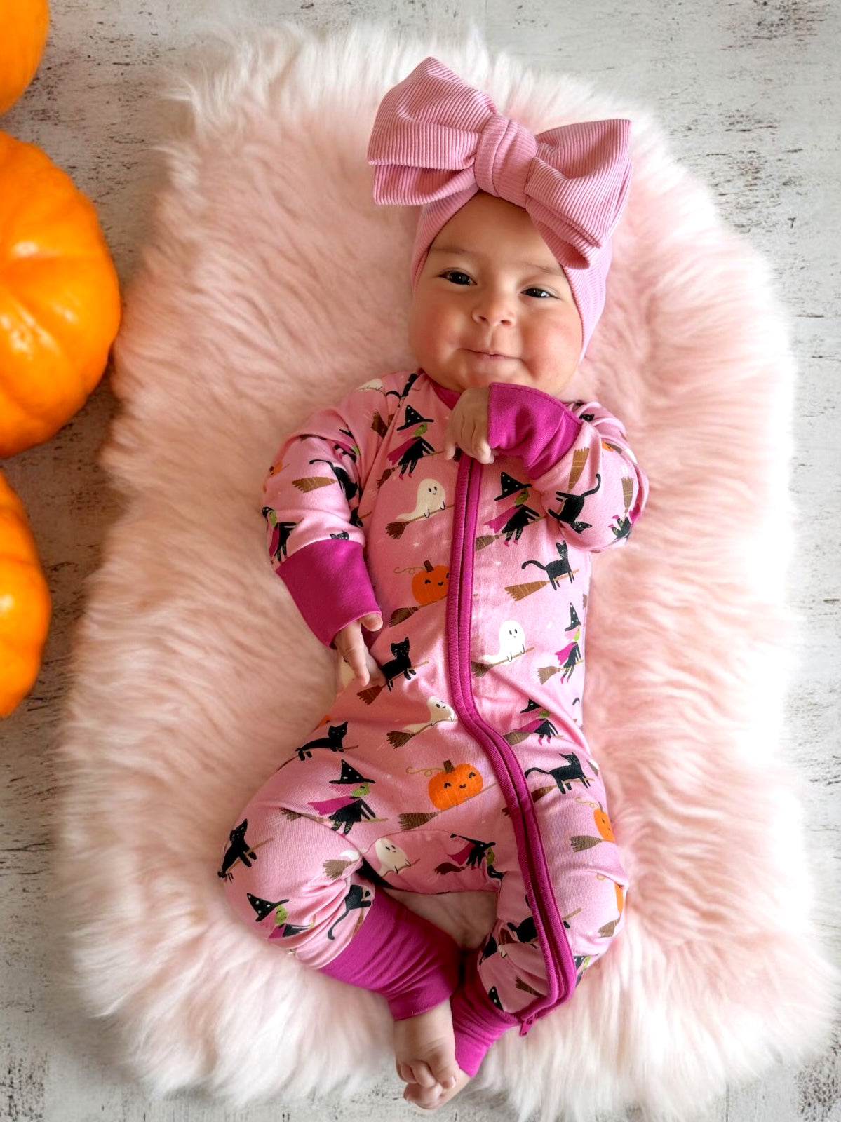 Smiling baby in a pink Halloween-themed onesie with a large bow, surrounded by pumpkins on a plush backdrop.