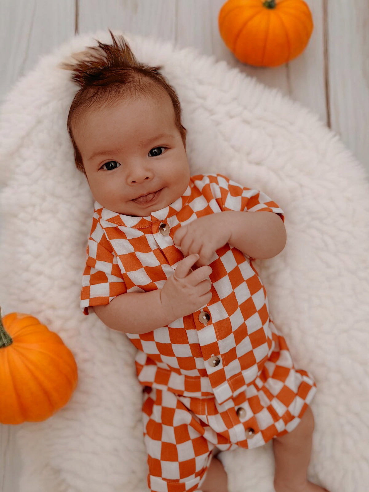 Smiling baby in an orange and white checkered outfit on a cozy blanket, surrounded by small pumpkins.