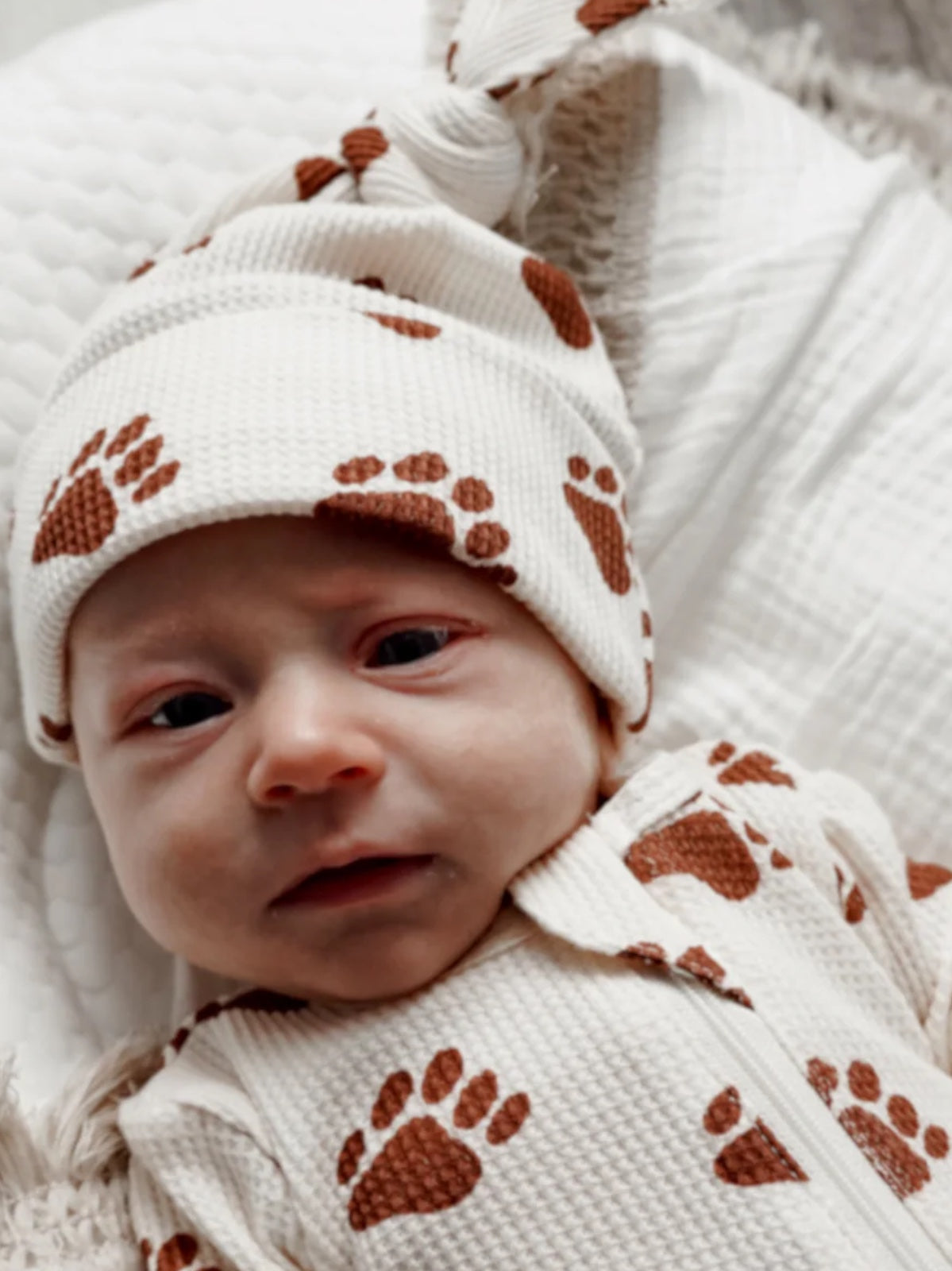 Baby wearing a cream outfit with brown paw prints and a matching hat, looking curiously at the camera.